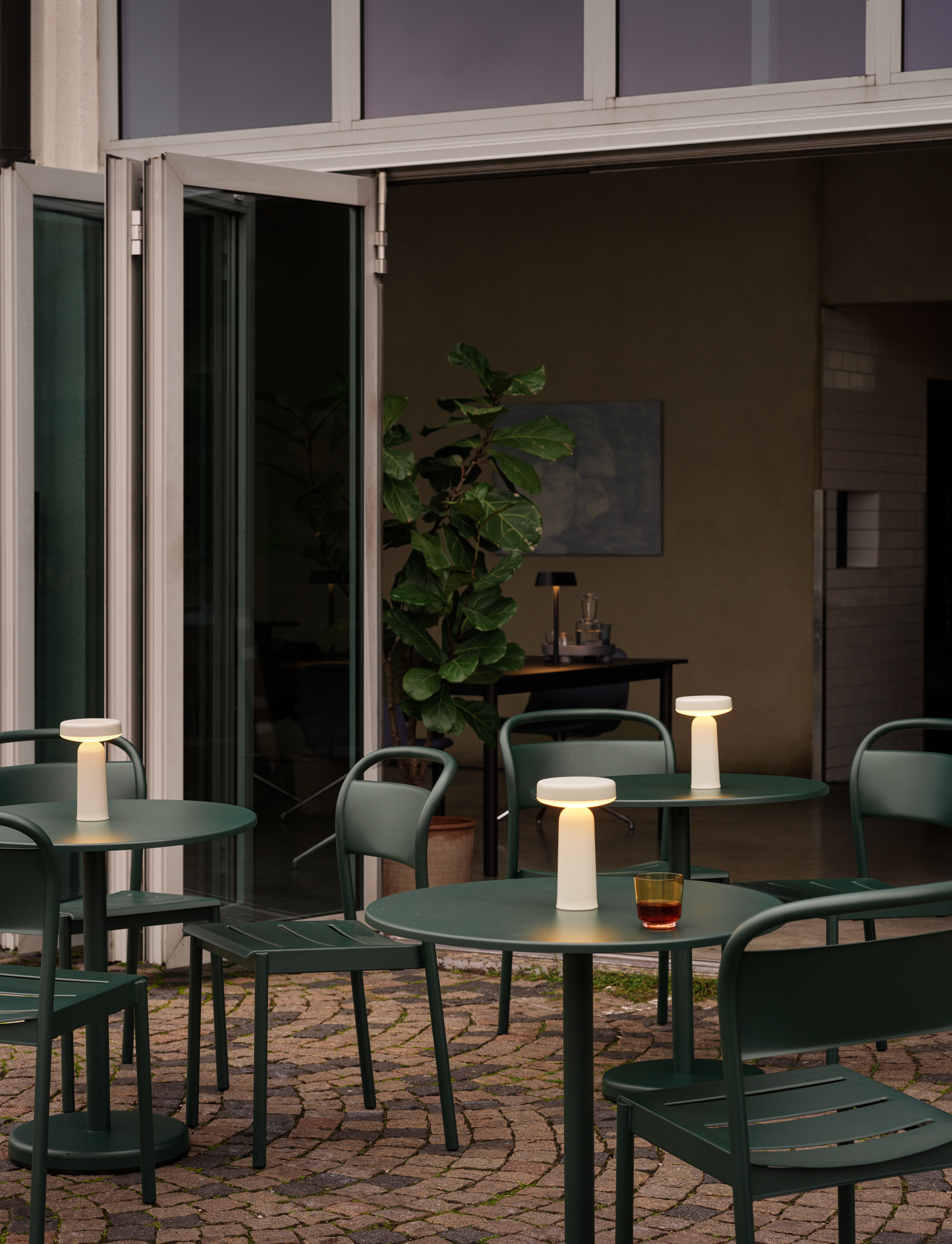 An outdoor café with dark green tables and chairs, each featuring a glowing Ease Lamp. The setting blends indoor and outdoor spaces with open glass doors and potted greenery.





