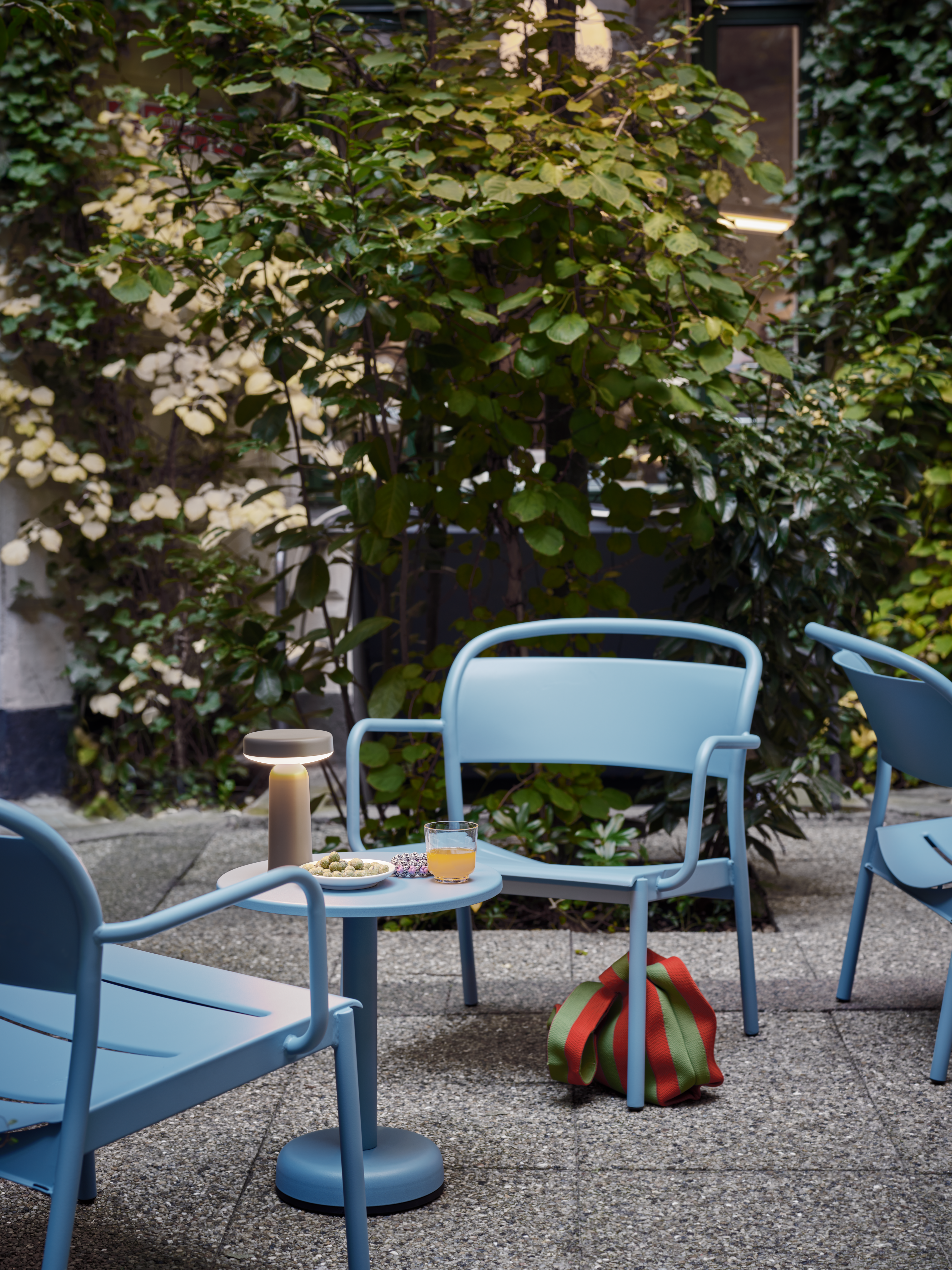 A cozy courtyard setting with light blue metal chairs and a small round table, illuminated by a soft-glowing Ease Lamp. A drink and snacks rest on the table.
