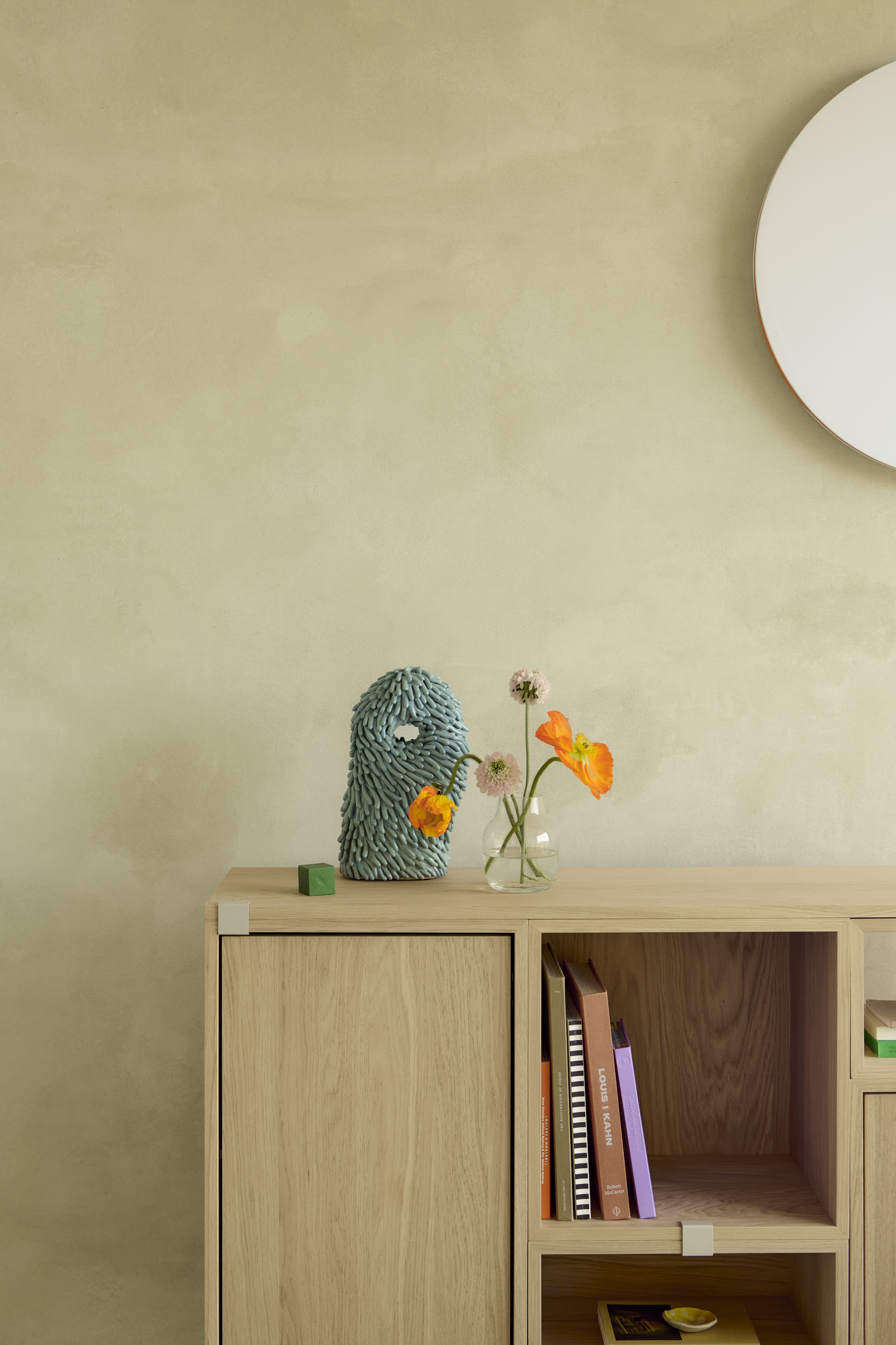 Stacked in oak is placed in a minimalistic room with brushed stone walls. A round wall lamp hangs above the storage system, casting a soft light that adds a subtle, elegant touch to the clean design.