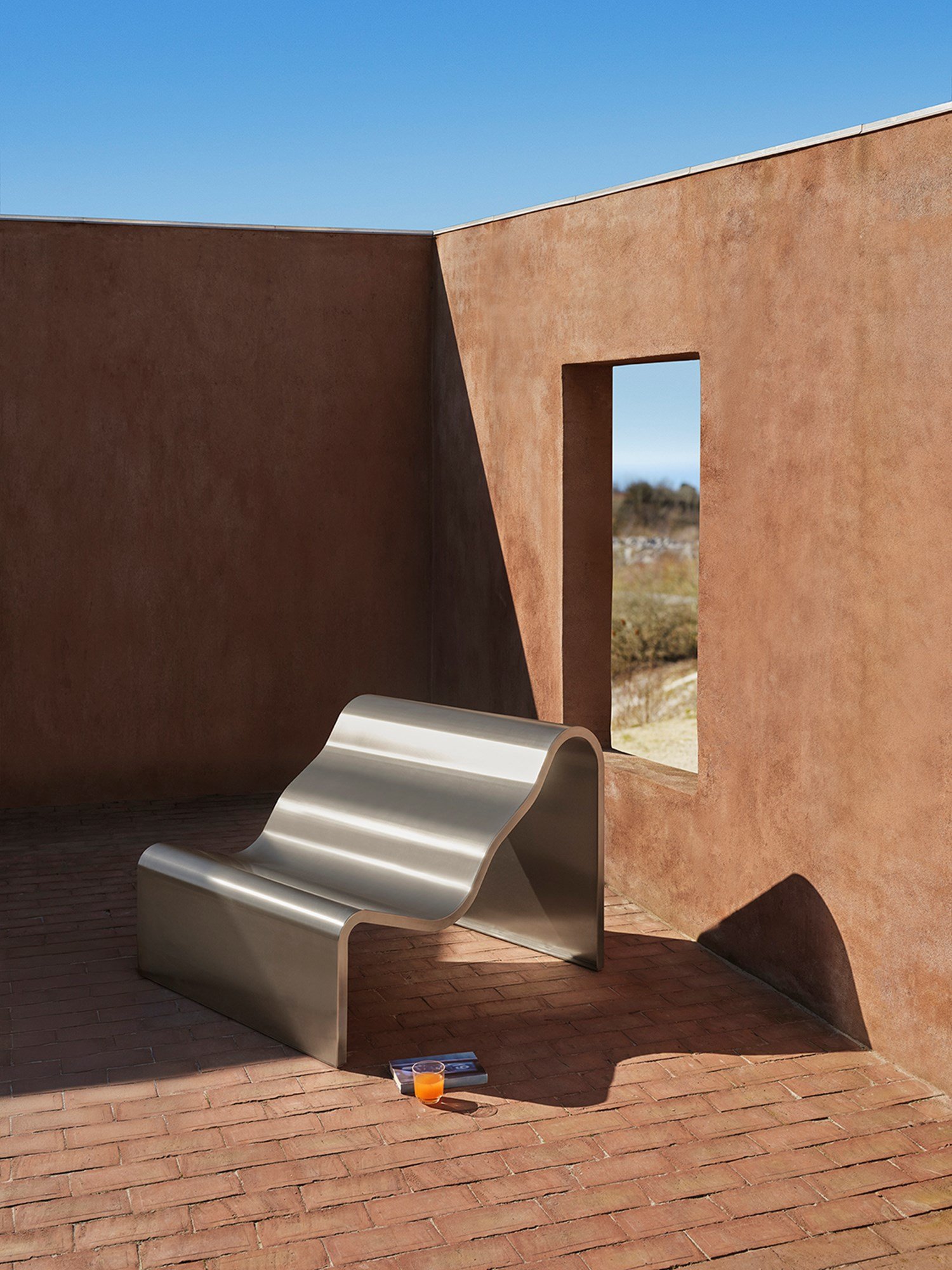 A architectural metal bench with a wavy, sculptural design is placed outdoor. A glass in a clear color is placed beside the bench.  A architectural metal bench with a wavy, sculptural design is placed outdoor. A glass in a clear color is placed beside the bench.