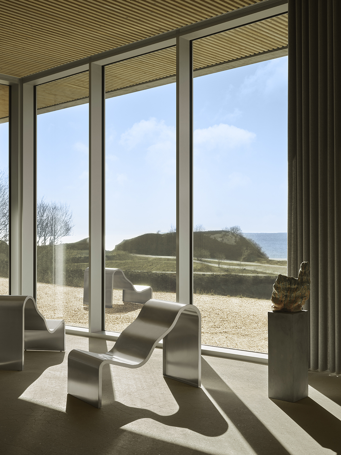 A architectural metal bench with a wavy, sculptural design is placed in front of a big window. 