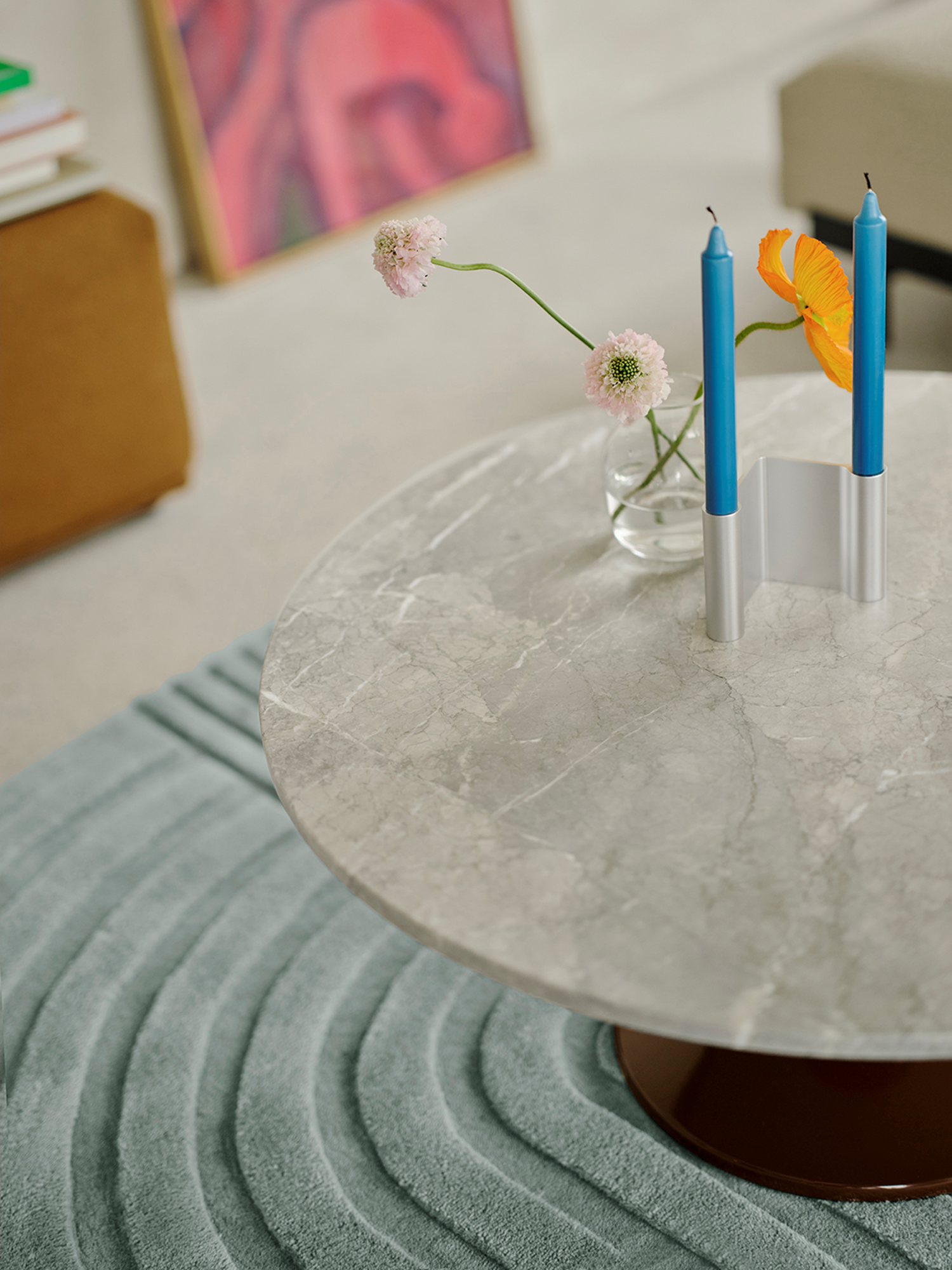 A close-up of a burgundy and marble coffee table, with a modern candle holder and a glass vase with white flowers placed on top. A soft light blue rug is positioned underneath the table, adding warmth and creating a cozy yet contemporary setting.