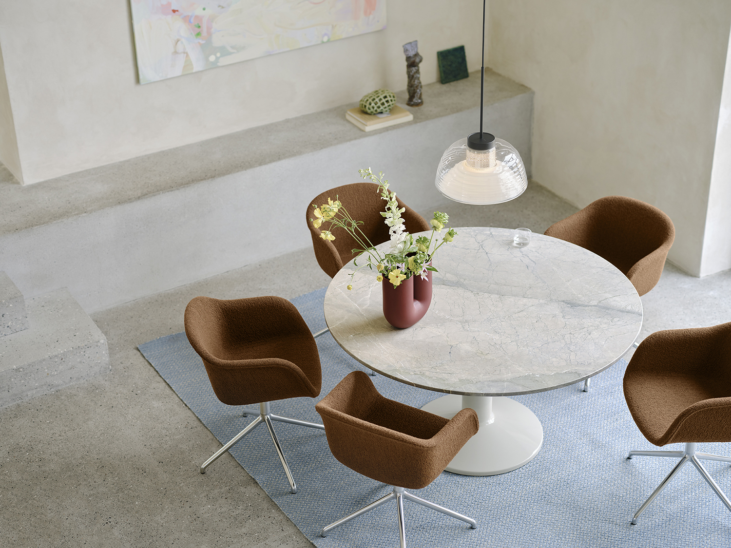 Seen from above, a round grey marble dining table sits in a minimalistic dining room with brushed stone walls and floors. Soft brown dining chairs surround the table, with a light blue rug beneath adding a pop of color. A glass pendant lamp hangs overhead, enhancing the serene and modern atmosphere.