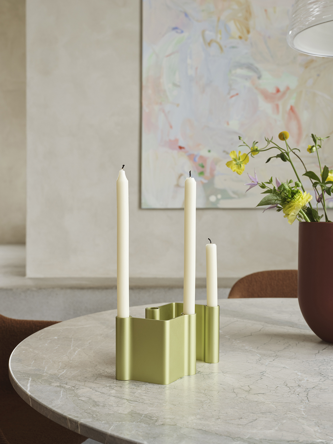 A modern candle holder in light green metal sits on a marble table, styled alongside a sculptural vase in a deep burgundy color for a striking, contemporary look.