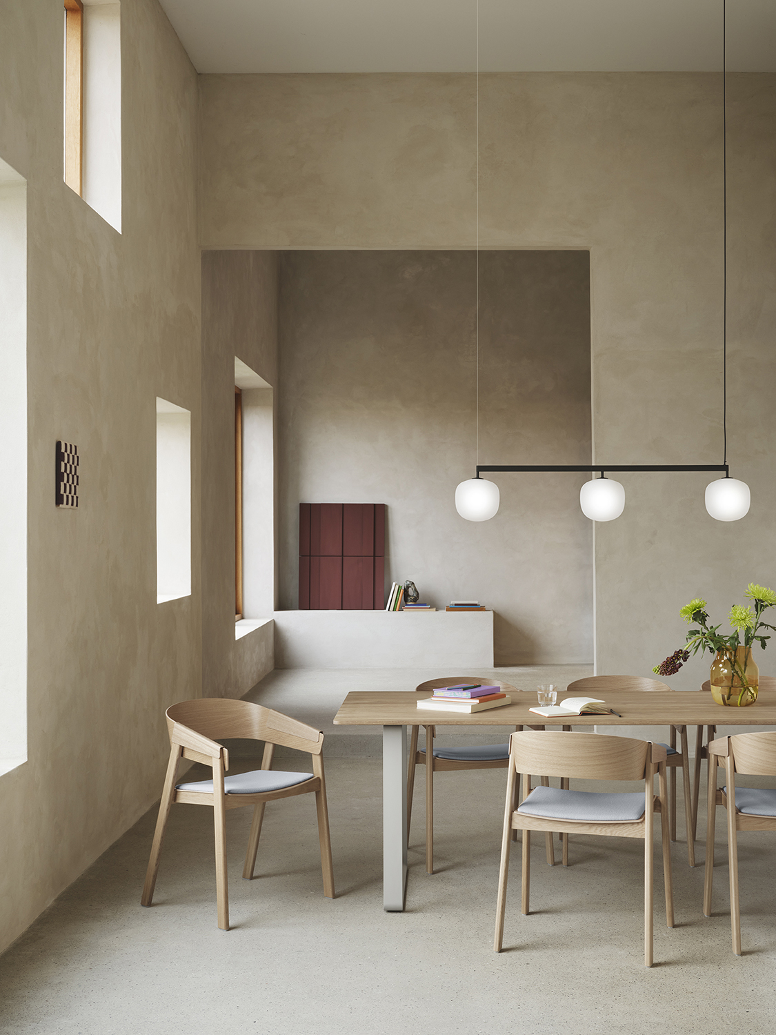 Modern wooden dining chairs are arranged around a wooden dining table in a minimalistic kitchen lounge area with brushed stone surfaces. A sleek, modern pendant lamp hangs above the table setting, enhancing the clean and natural aesthetic.