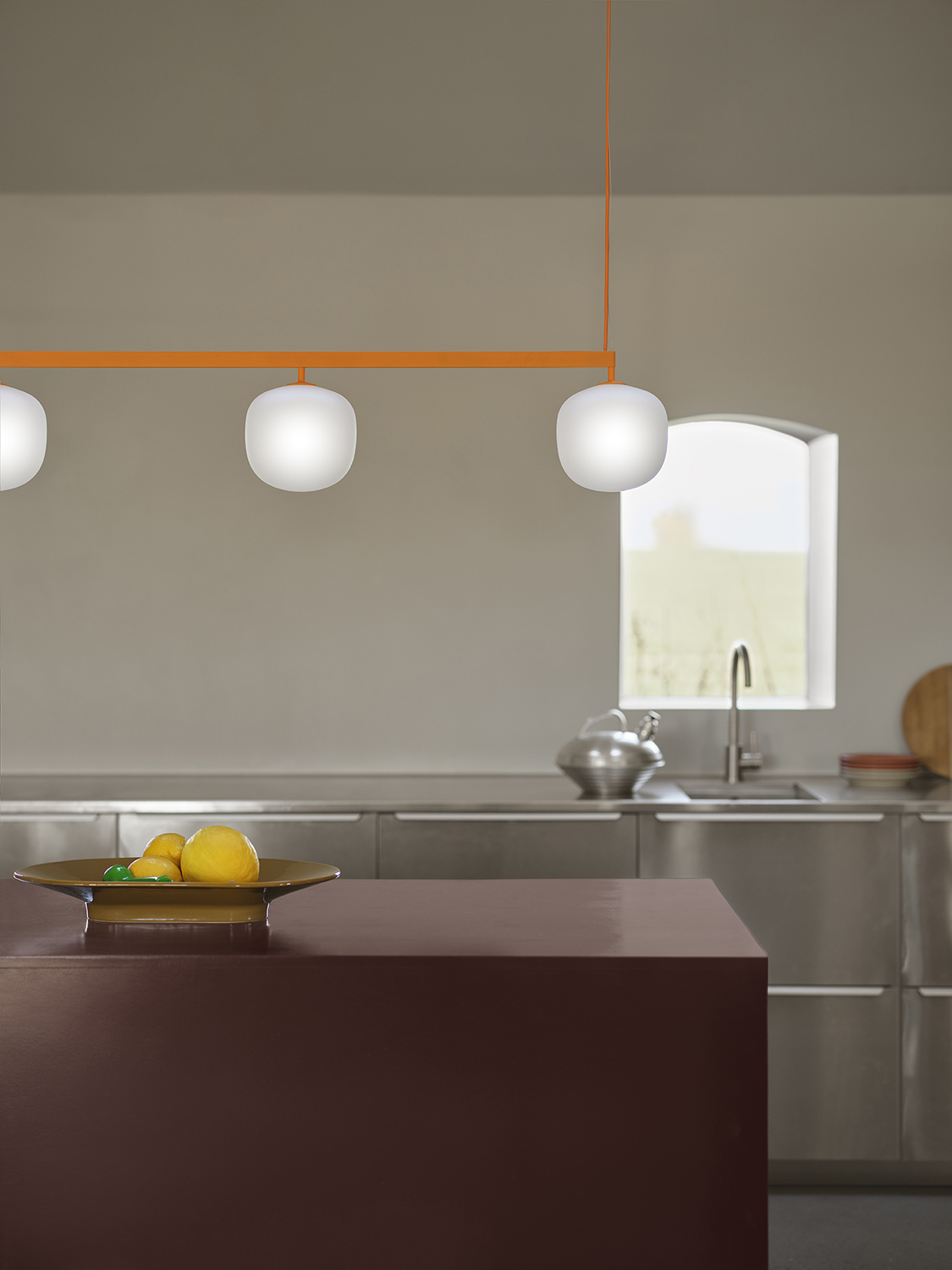 An orange pendant lamp hangs above a dark burgundy kitchen table, where a brown ceramic bowl is placed, adding warmth and contrast to the modern setting.