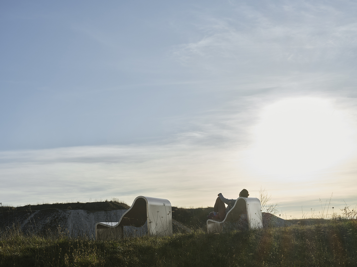 A metallic bench with a soft, architectural design, positioned outdoors on a grassy area and bathed in sunlight.
