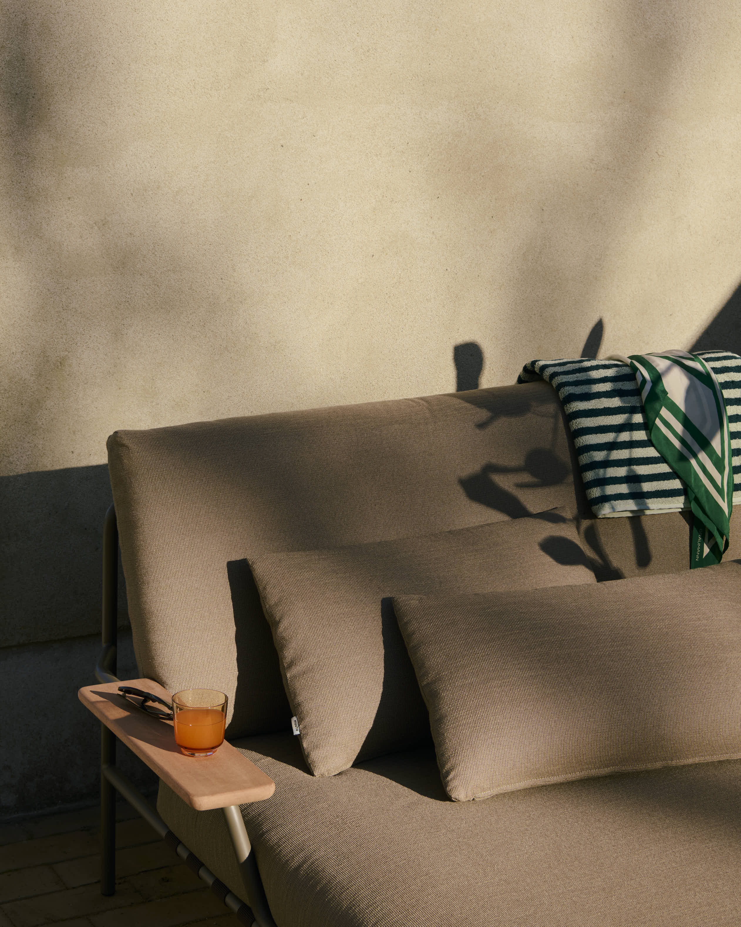A close-up of a modern, sleek lounge bed in a dark sand-colored fabric with an armrest, positioned outdoors and bathed in the warm light of the afternoon sun.