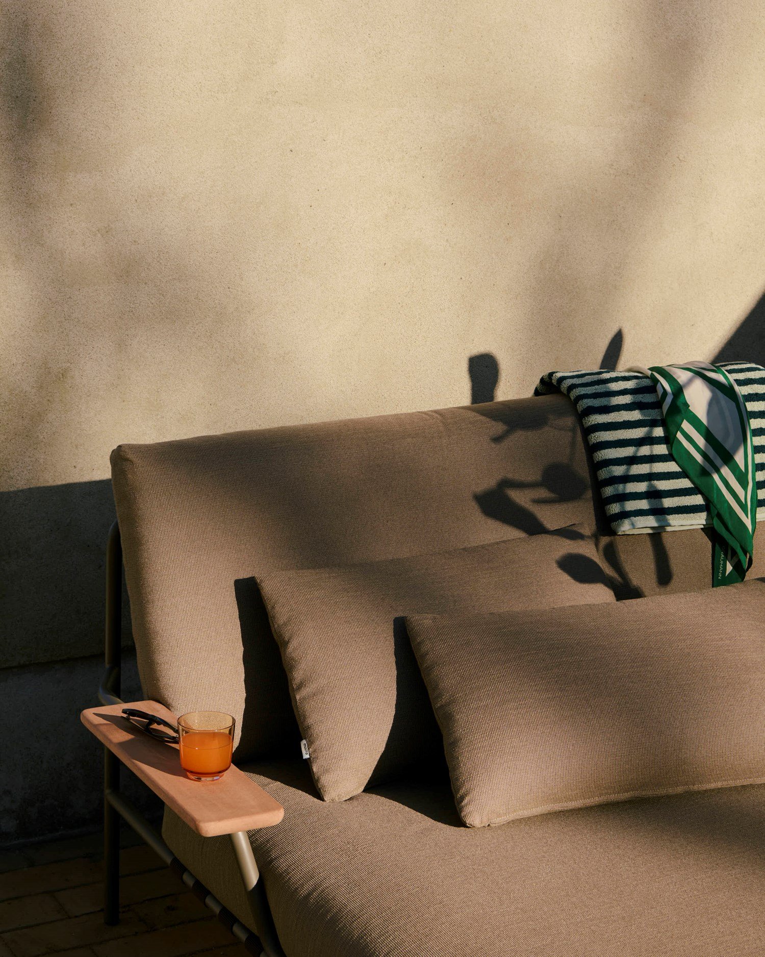 A close-up of a modern, sleek lounge bed in a dark sand-colored fabric with an armrest, positioned outdoors and bathed in the warm light of the afternoon sun. A close-up of a modern, sleek lounge bed in a dark sand-colored fabric with an armrest, positioned outdoors and bathed in the warm light of the afternoon sun.