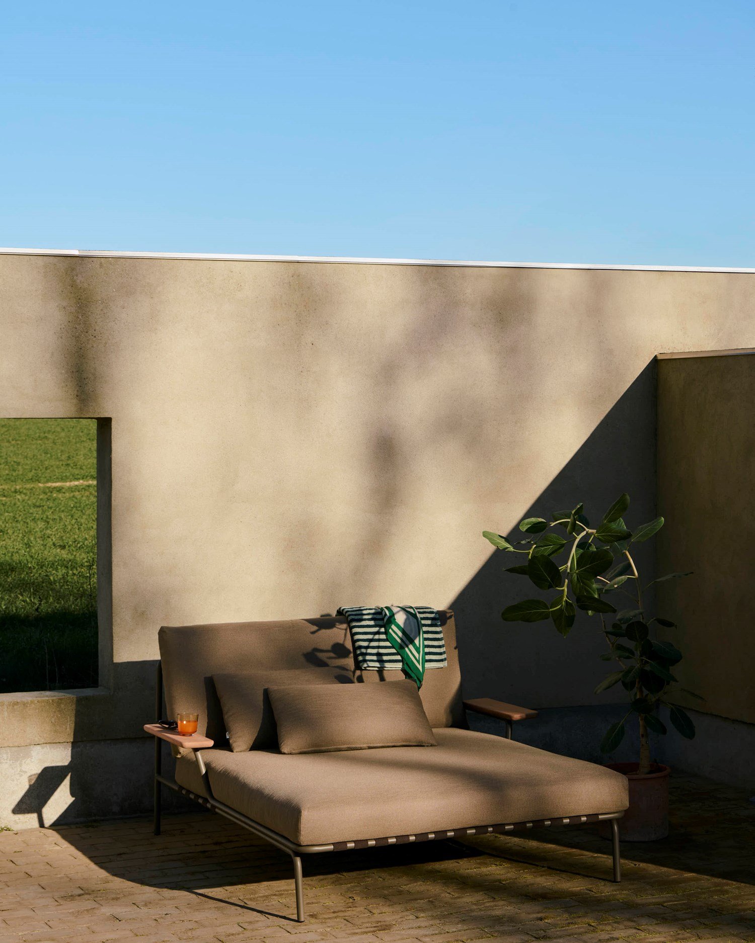 A modern, sleek lounge bed in a dark sand-colored fabric with an armrest, positioned outdoors and bathed in the warm light of the afternoon sun. A modern, sleek lounge bed in a dark sand-colored fabric with an armrest, positioned outdoors and bathed in the warm light of the afternoon sun.