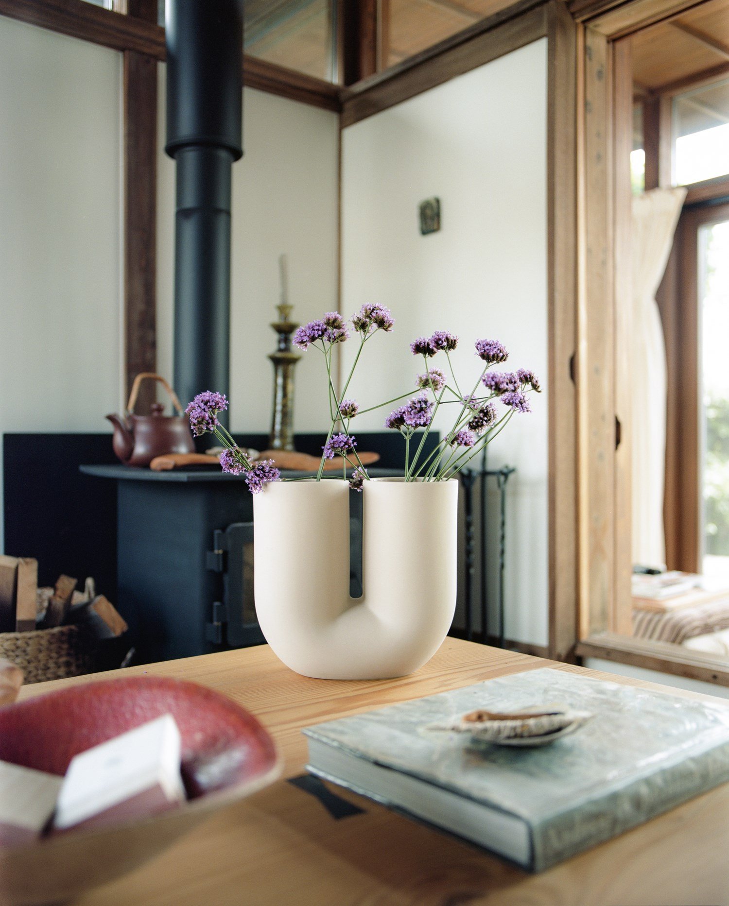 Kink Vase in the color sand with purple flowers on a table in a rustic room with a stove.
 Kink Vase in the color sand with purple flowers on a table in a rustic room with a stove.