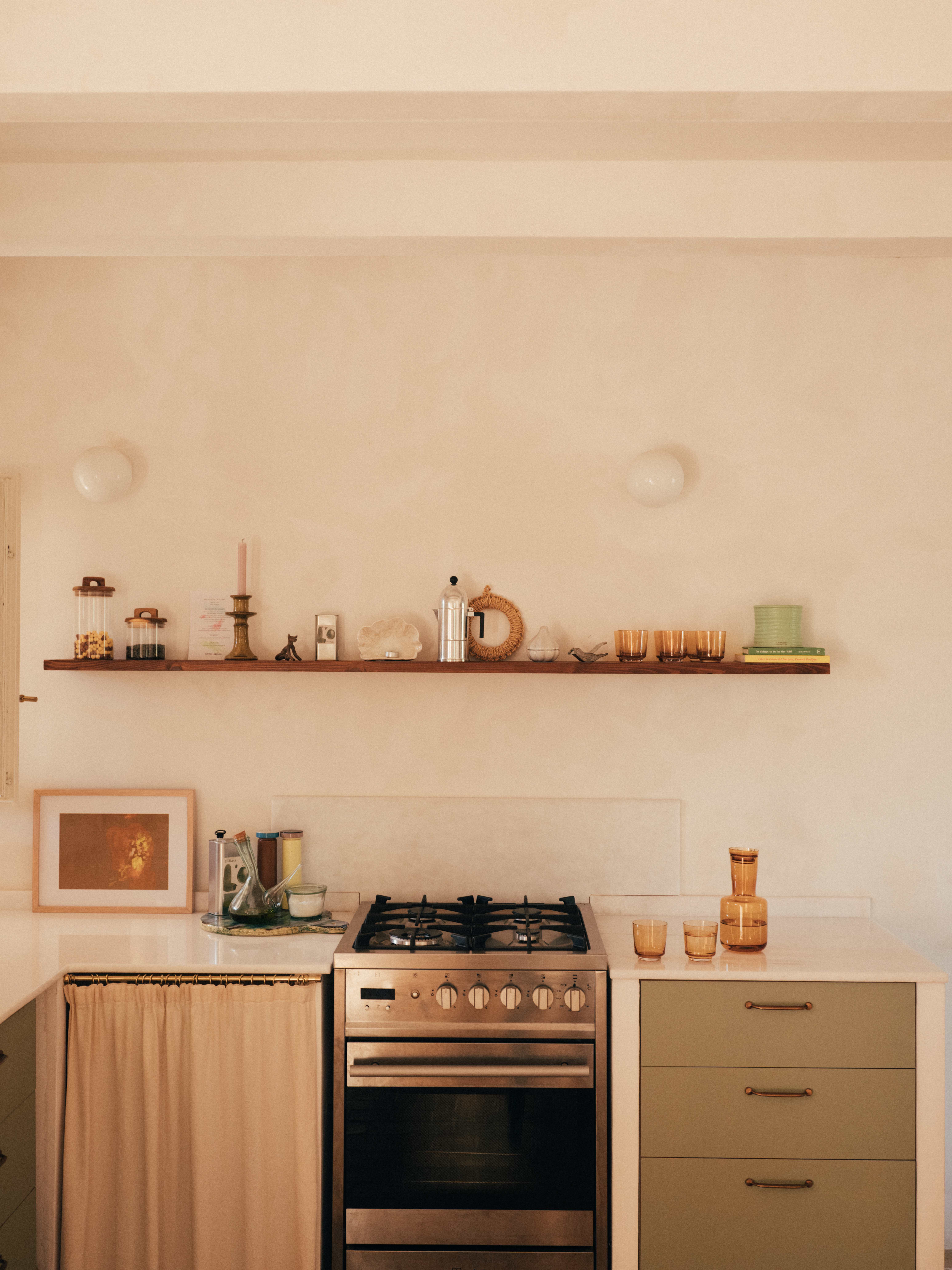 A minimalist kitchen with rustic charm – A warm, neutral-toned kitchen featuring a stainless steel gas stove centered between sage green cabinets with brass handles and a beige curtain covering storage space. Above, a floating wooden shelf holds ceramics, glassware, and decorative objects, blending functionality with a soft Mediterranean aesthetic.