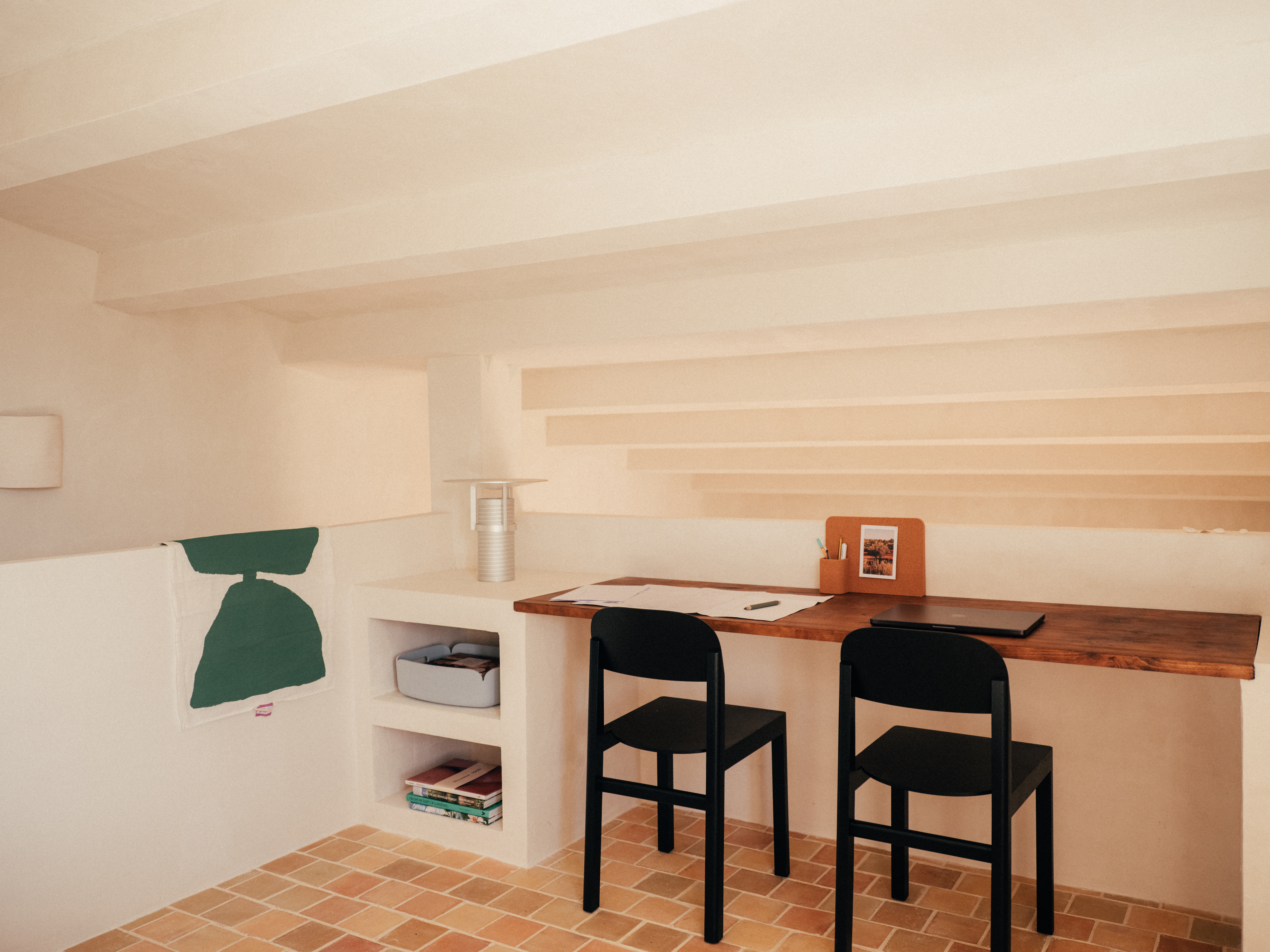 Close-up of a cozy desk setup – A detailed look at a wooden workspace with a sculptural Set Lamp, cork organizer, and a black chair. Below, open built-in shelves store books and a soft gray storage bin, seamlessly blending practicality with the rustic, minimalist decor.