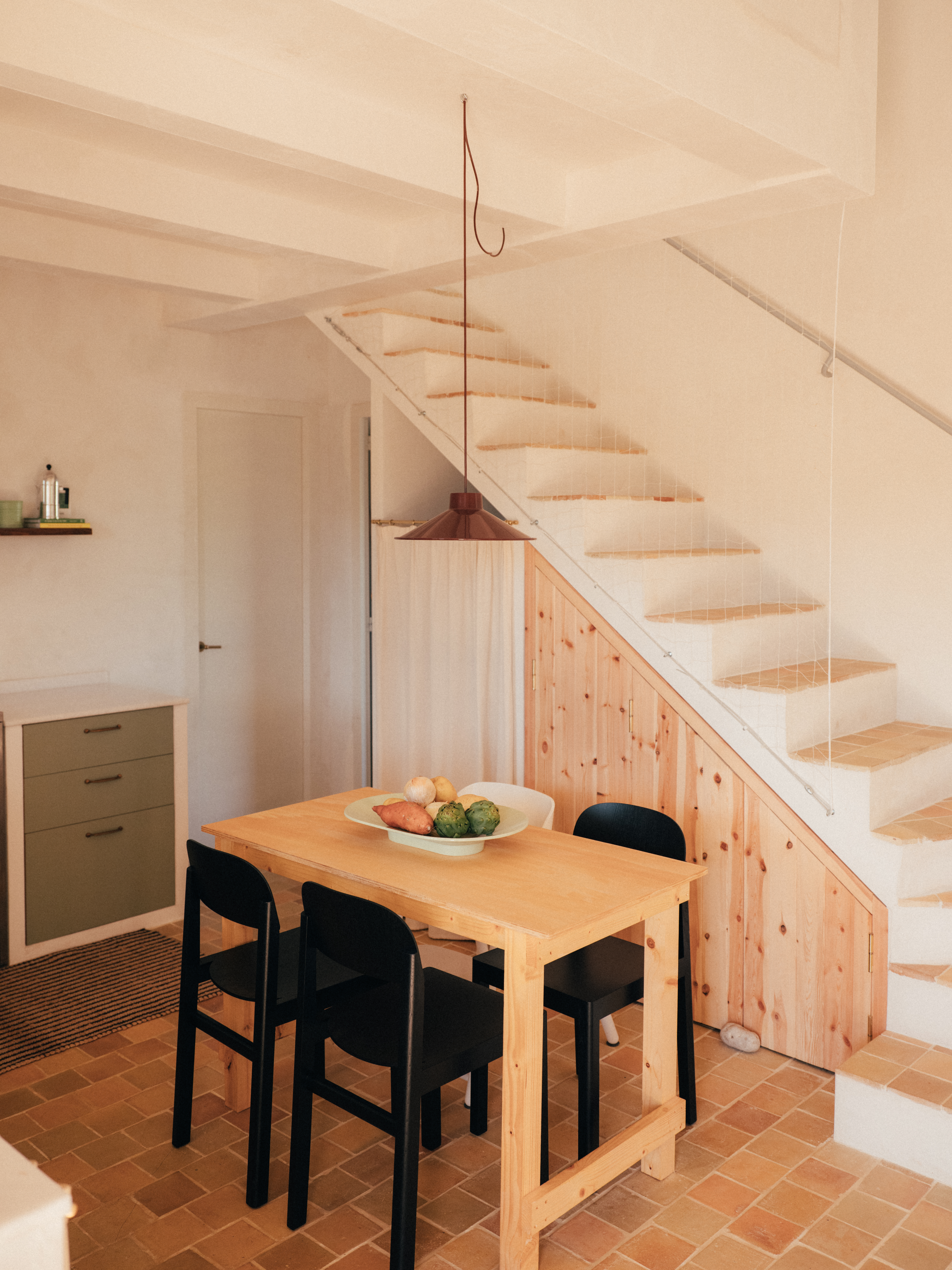 A warm and inviting kitchen-dining space – A natural wood dining table with black chairs sits under a terracotta pendant lamp. A bowl of fresh vegetables adds color to the rustic setting. In the background, a staircase with wooden paneling and white railings leads to the upper level, complementing the home’s earthy tones.