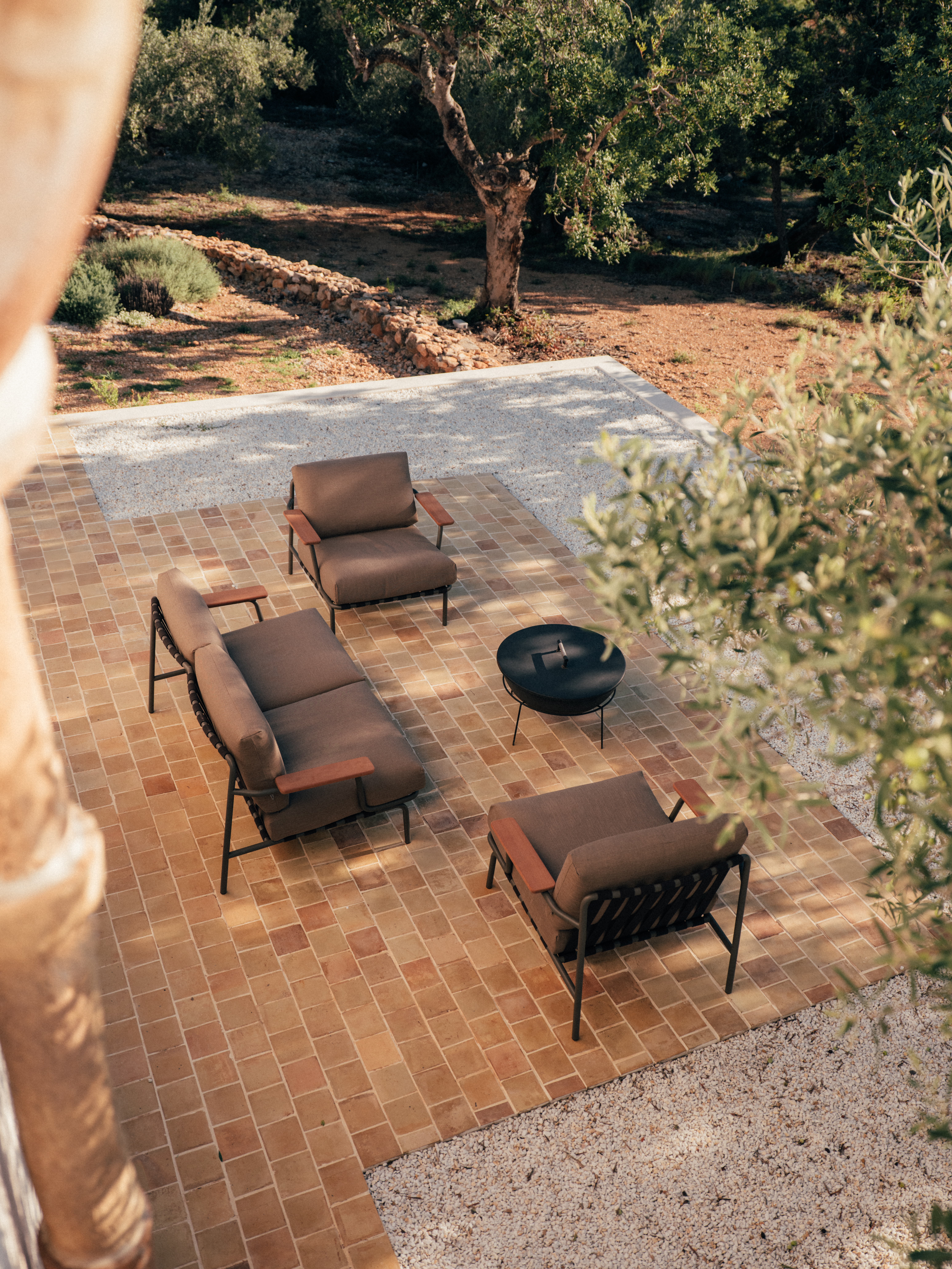 Cozy outdoor seating in a rustic landscape – A sunlit patio with a Settle Sofas two matching armchairs, and a black fire pit. The furniture has a modern design with black metal frames and wooden armrests. The warm terracotta tiles contrast with the surrounding gravel, olive trees, and a stone wall in the background.