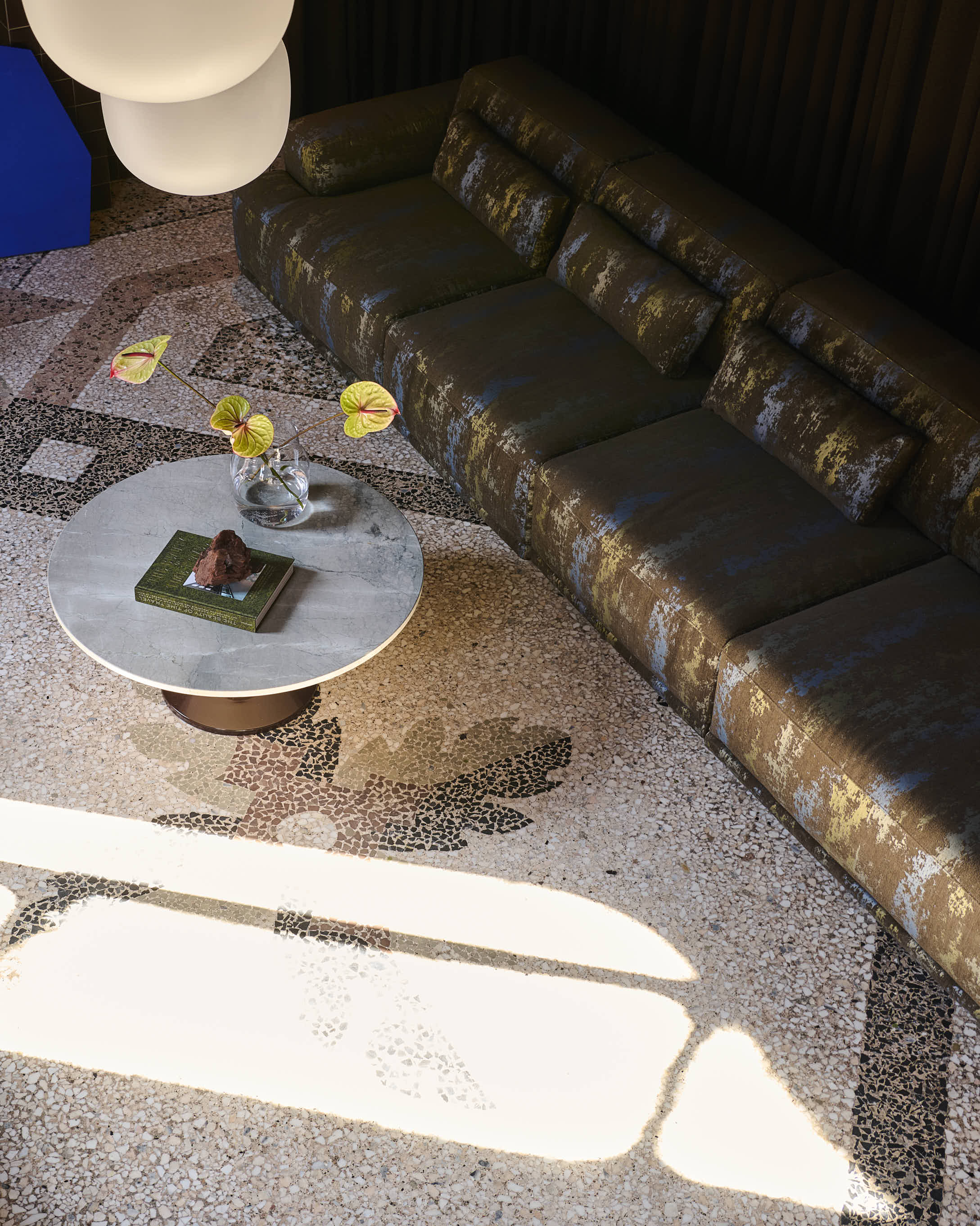 A close-up of a room seen from above. A ray of sunlight casts shadows on a stone mosaic floor. A marble sofa table is placed in the room, accompanied by a dark patterned sofa. A vase with flowers and a coffee table book sits on the table.