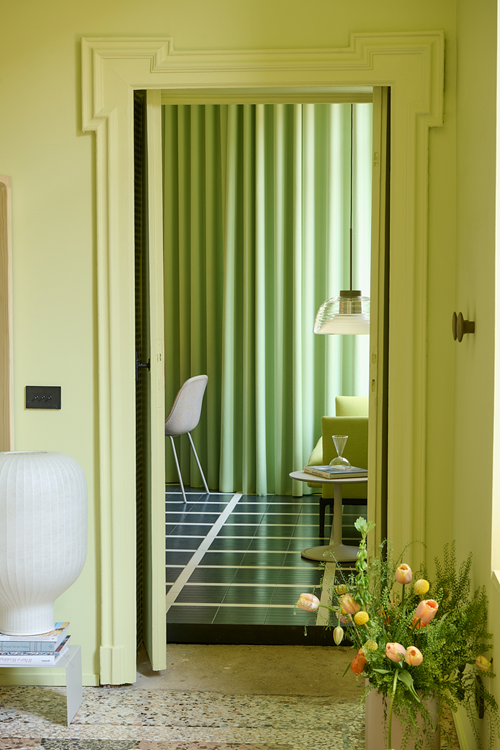 A light green room. An open door reveals a glimpse of another space with light green curtains, styled with a glass pendant lamp hanging above a table and a chair.