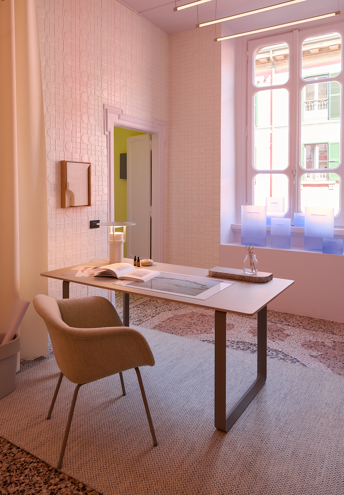 A light, bright dusty pink room styled with a soft chair and a modern wooden table. An aluminium table lamp is placed on the table, alongside a clear minimalist vase and a book. Light blue vases in frosted glass, at varying heights, are placed in the window.