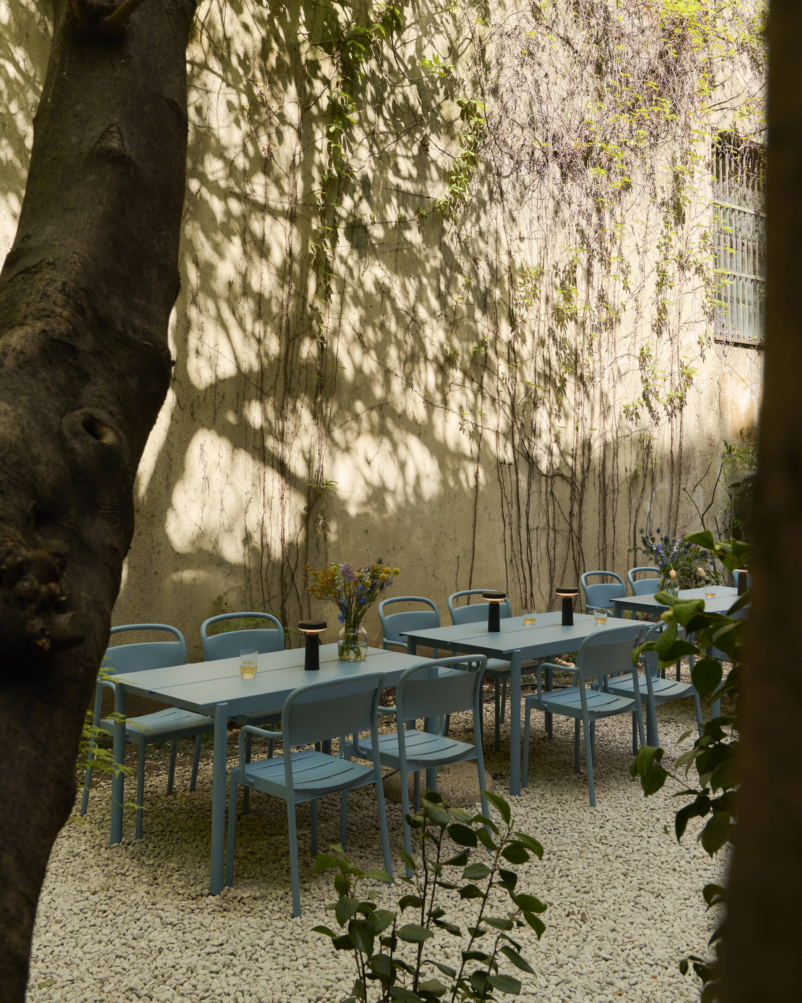 In a cozy courtyard, some outdoor tables and chairs are placed in the shade. The table setting is styled with vases of flowers and table lamps. Trees and flowers surround the setting.