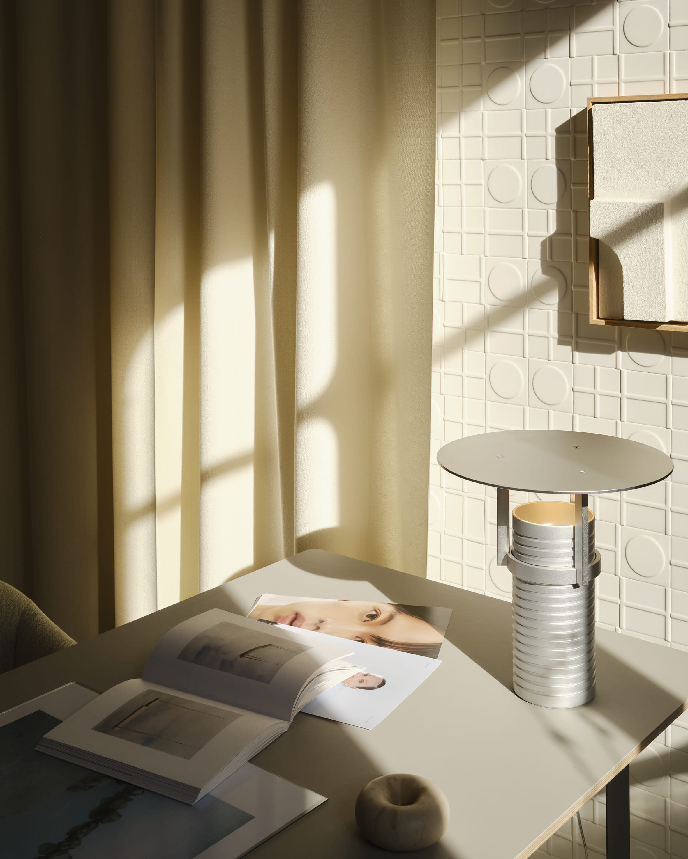 A close-up of a modern aluminum table lamp placed on a table. Rays of sunlight are shining on the table setting.
