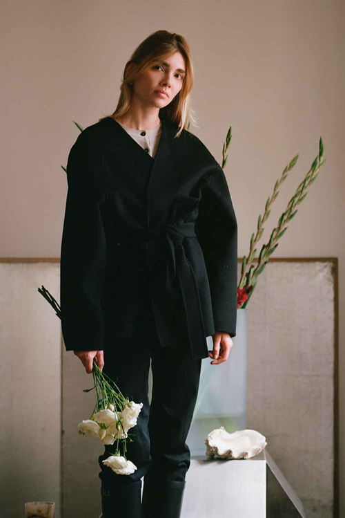 Nina stands holding a bouquet of white flowers in a black oversized jacket. Behind her, a glass vase with tall stems of greenery is displayed against a soft, neutral backdrop.