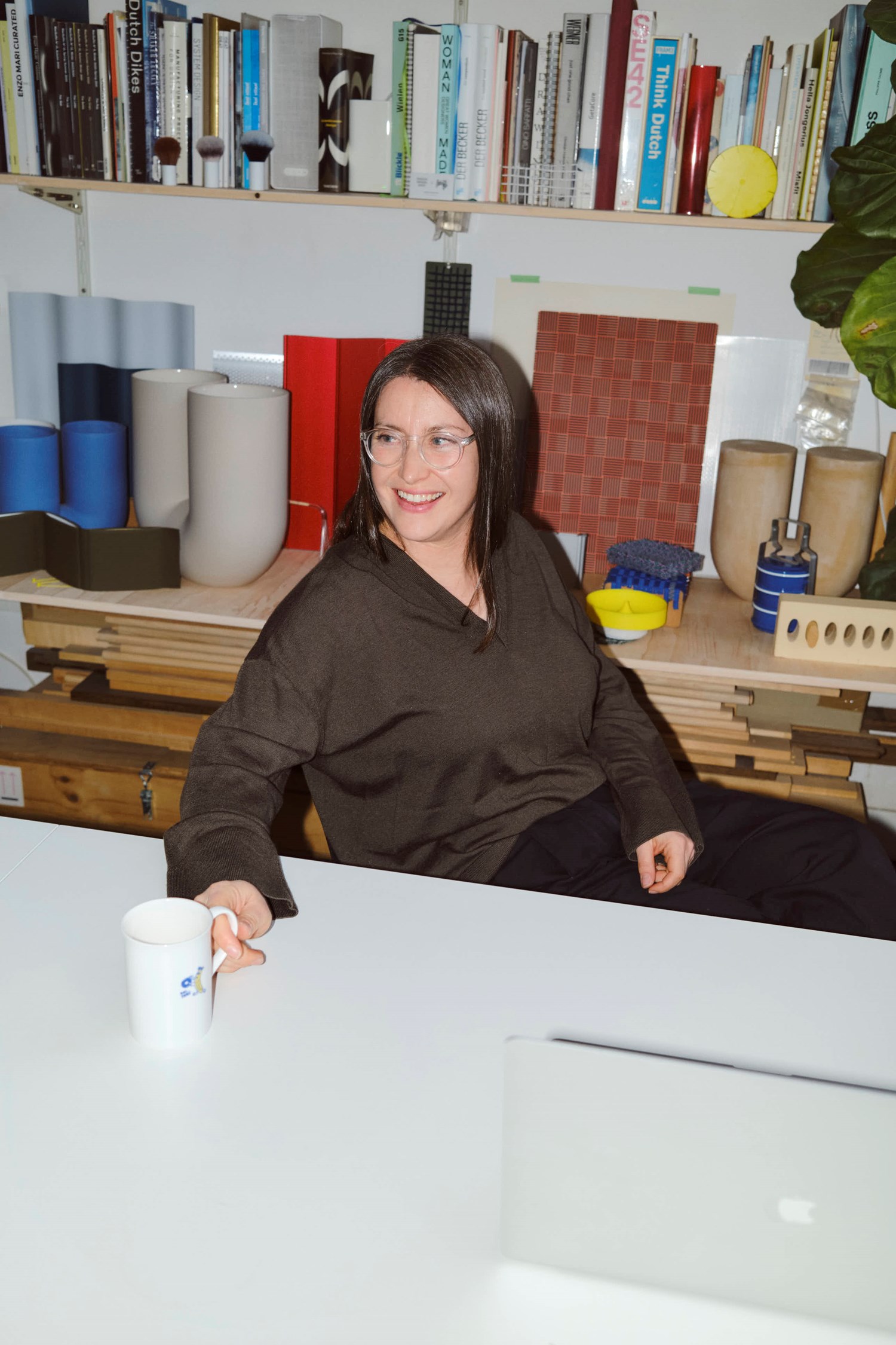Rachel Griffin smiles while seated at a white desk in her studio, holding a white ceramic mug. Behind her, several Kink Vases in matte blue and neutral tones are displayed among material samples, tools, and a large collection of design books. Rachel Griffin smiles while seated at a white desk in her studio, holding a white ceramic mug. Behind her, several Kink Vases in matte blue and neutral tones are displayed among material samples, tools, and a large collection of design books.