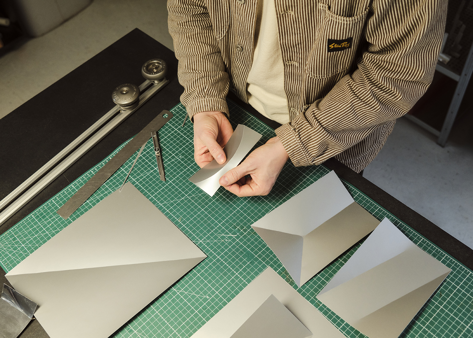 Close-up of a Phil’s hands gently folding a curved metal sheet above a green cutting mat. Surrounded by various sharply creased metal forms, a precision knife, and measuring tools.

