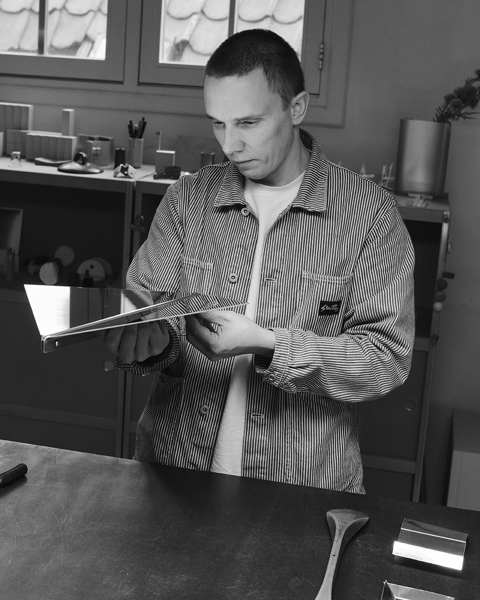 Phil in a striped jacket stands at a large worktable in a modern design studio, carefully examining a sheet of folded reflective metal.