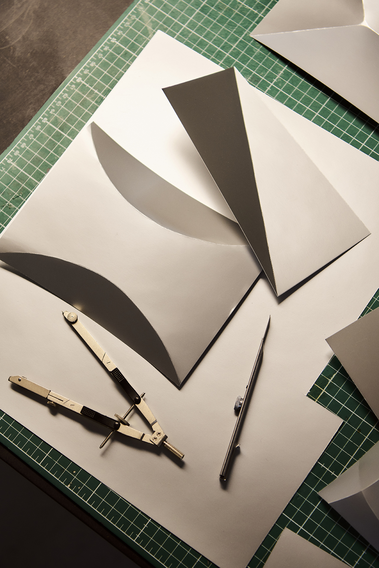 A green cutting mat displays folded and curved silver paper or metal prototypes, precision compasses, and a hobby knife. The scene is lit warmly, emphasizing the geometric purity and design refinement of sculptural studies or product development mock-ups.

