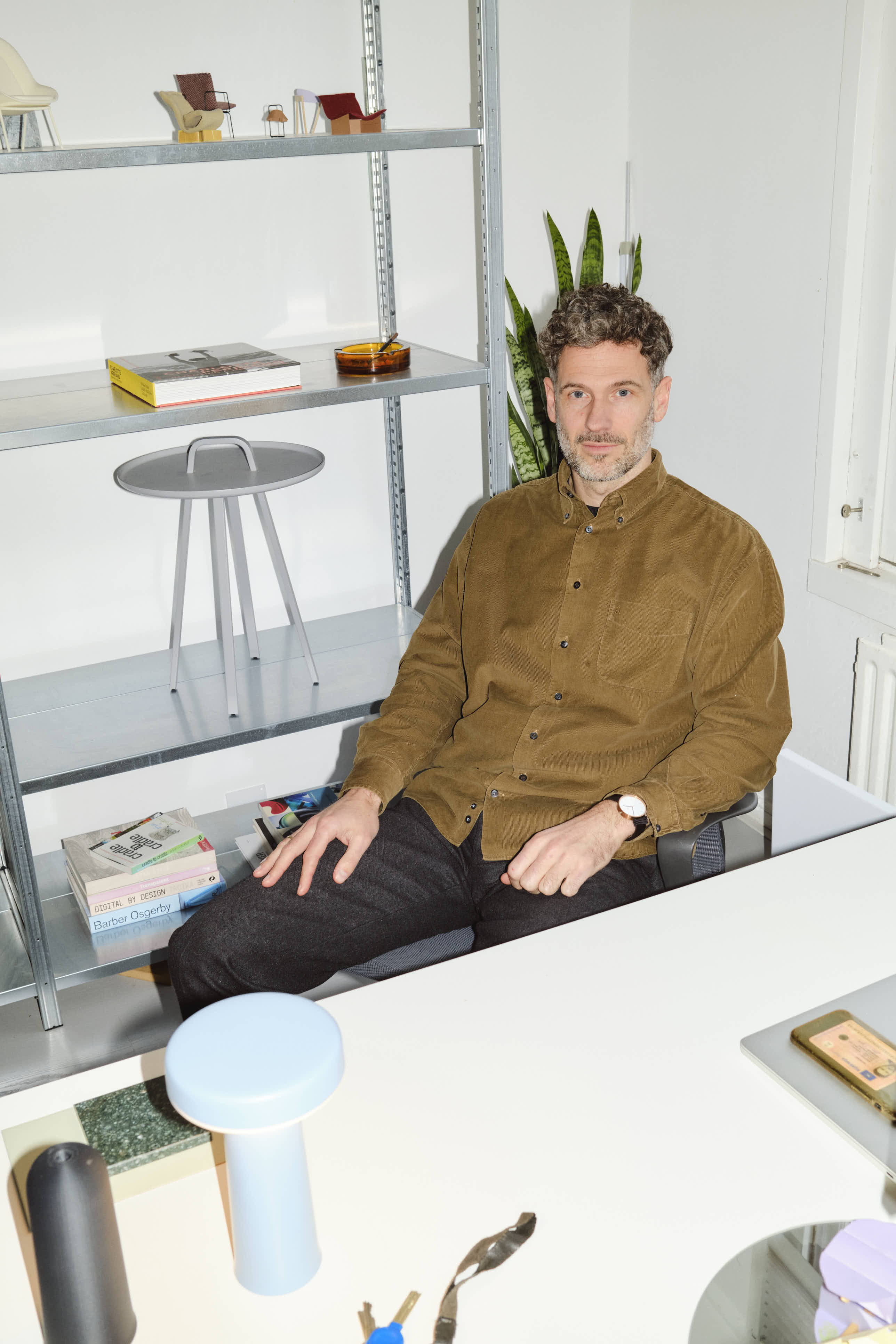 Johan in a brown shirt sits at a desk in a design studio, with a light blue Muuto Ease portable lamp in front of him and shelves filled with books and objects behind him.
