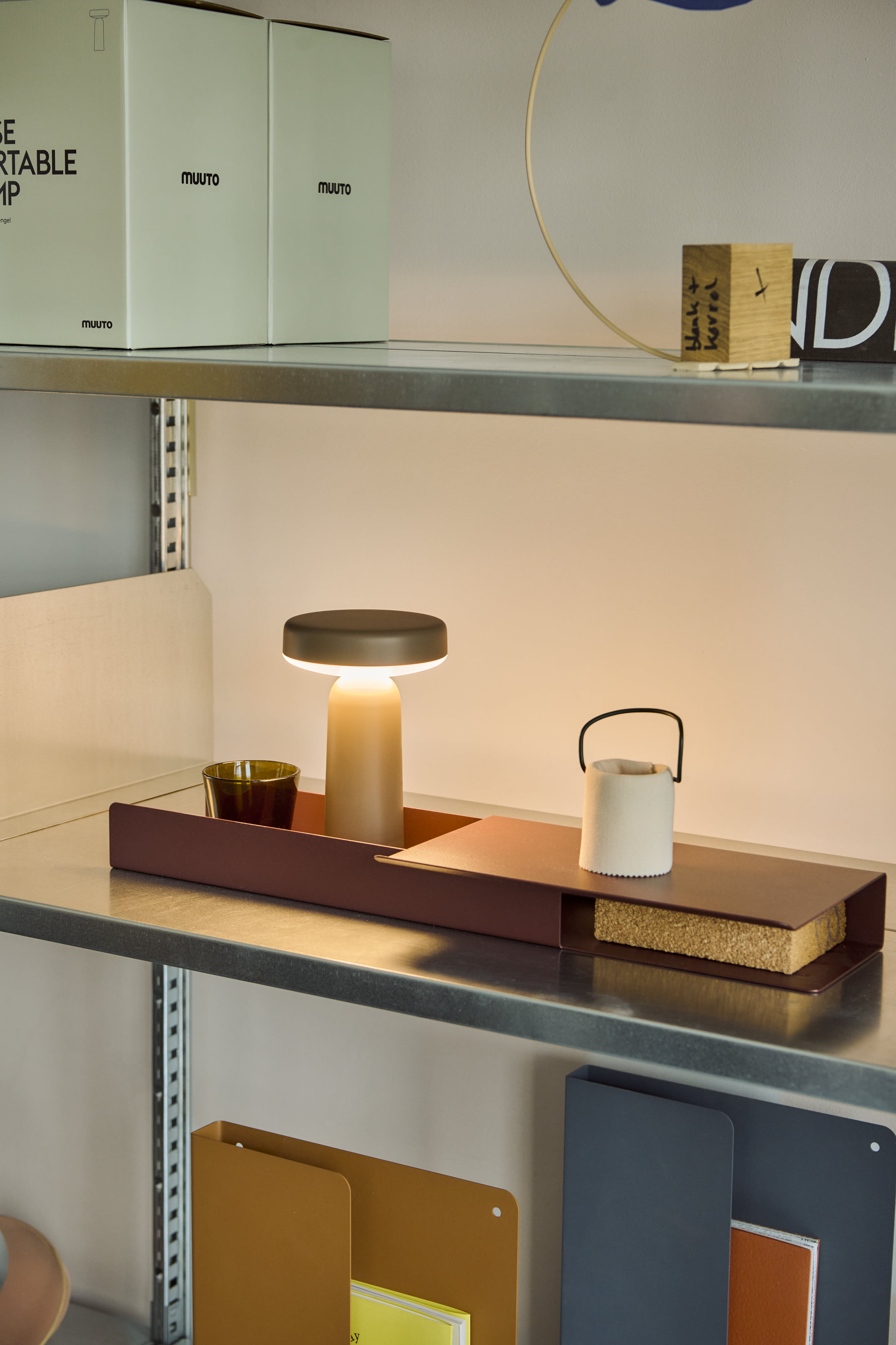 A taupe Muuto Ease portable lamp emits a warm glow on a metal shelf, alongside a glass cup, a small white lantern, and neatly arranged office supplies.
