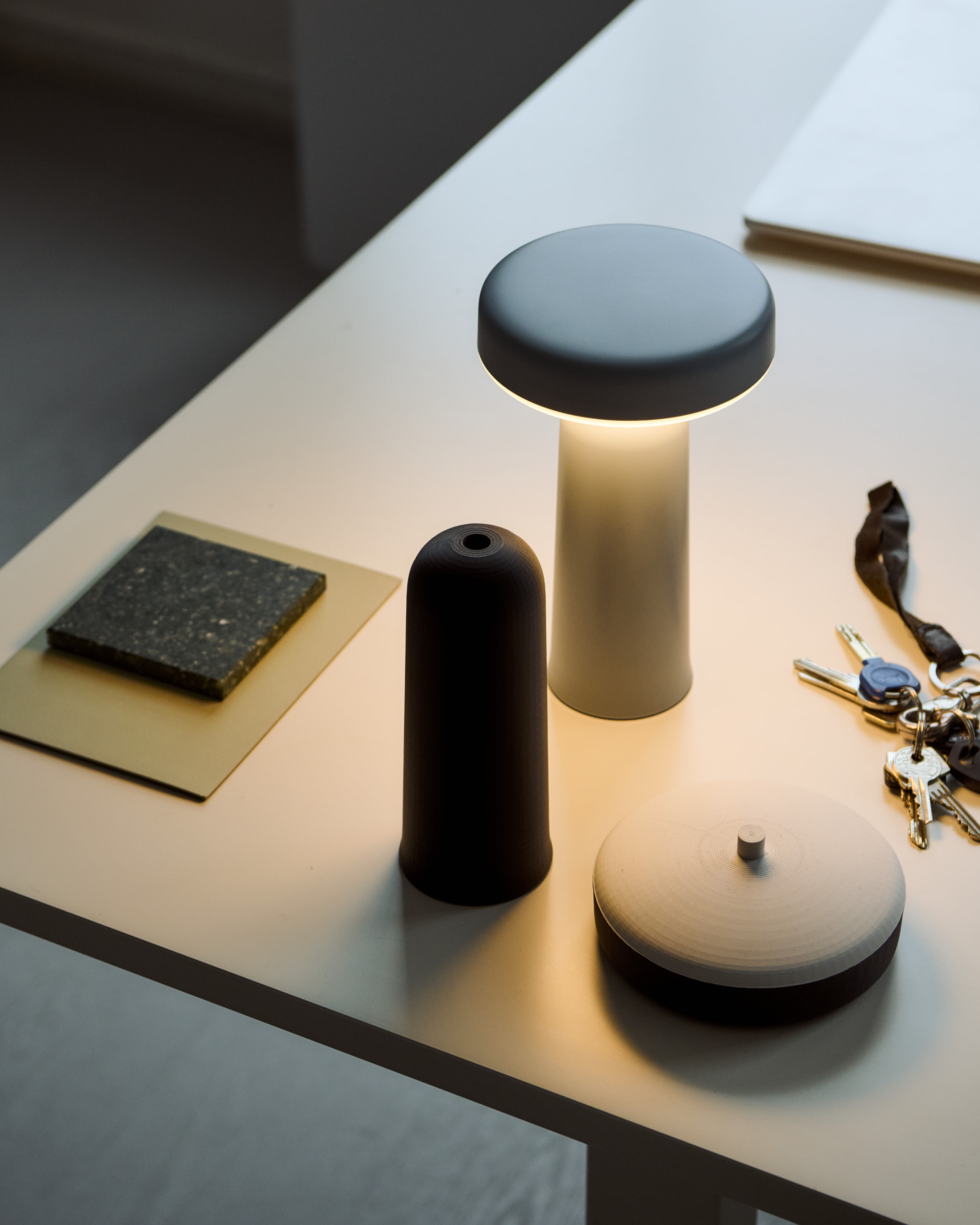 A lit Muuto Ease portable lamp with a blue top and white base sits on a desk, accompanied by a black lamp cover, keys, and textured material samples.