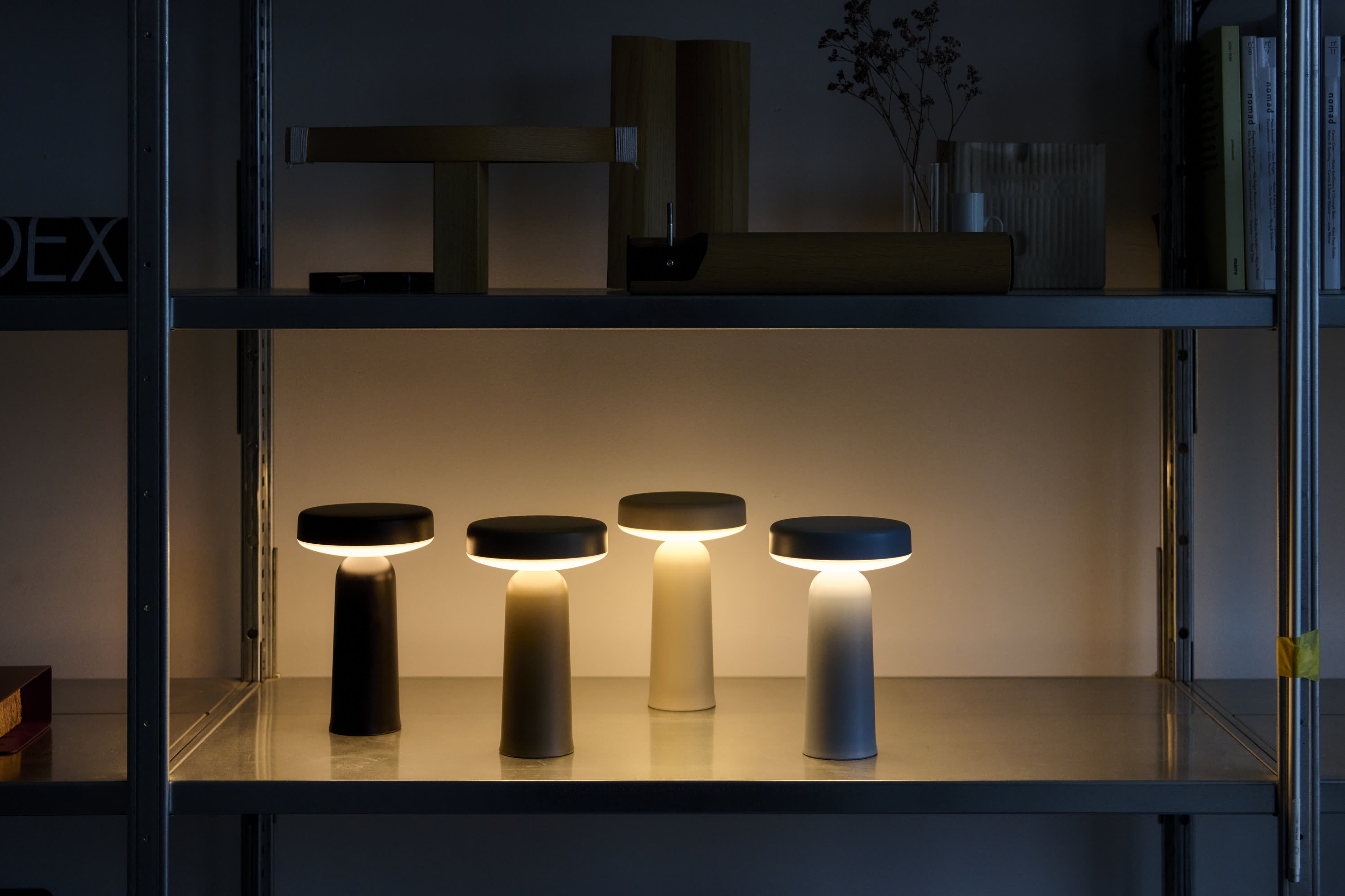 Four Muuto Ease portable lamps glowing in a dimly lit room, placed on a metal shelf among wooden and metal objects, creating a warm ambiance.