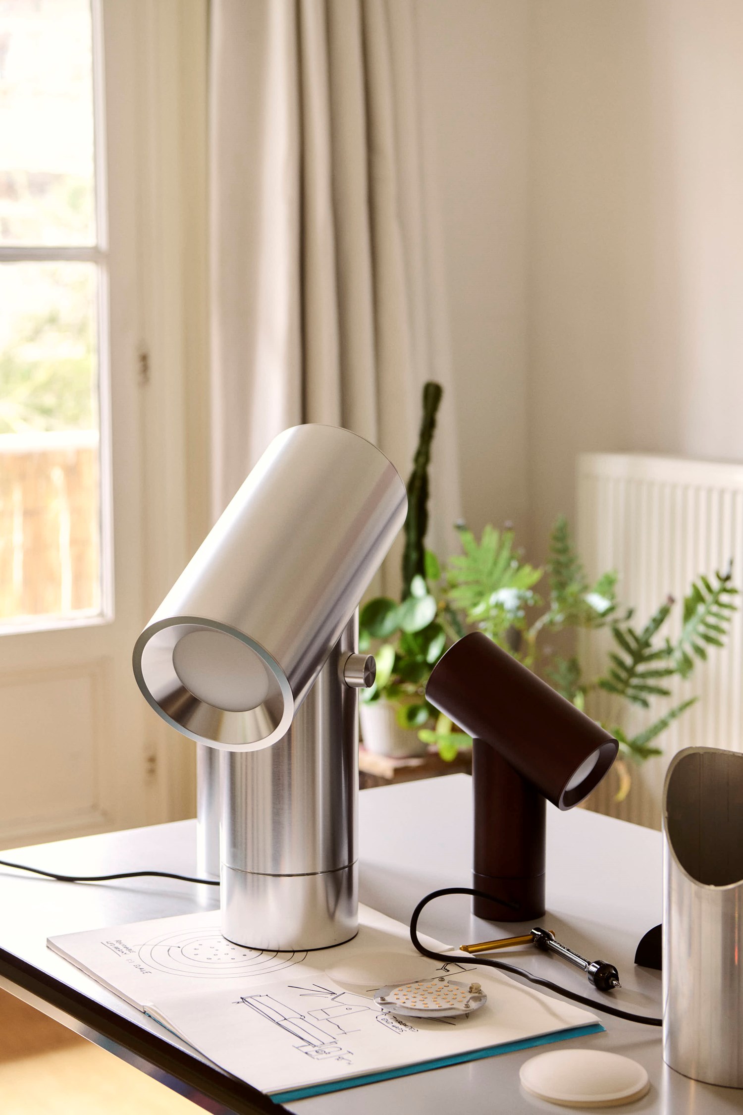 A Modern setting with silver and black Beam table lamps on a desk with papers, near a window and indoor plants. A Modern setting with silver and black Beam table lamps on a desk with papers, near a window and indoor plants.