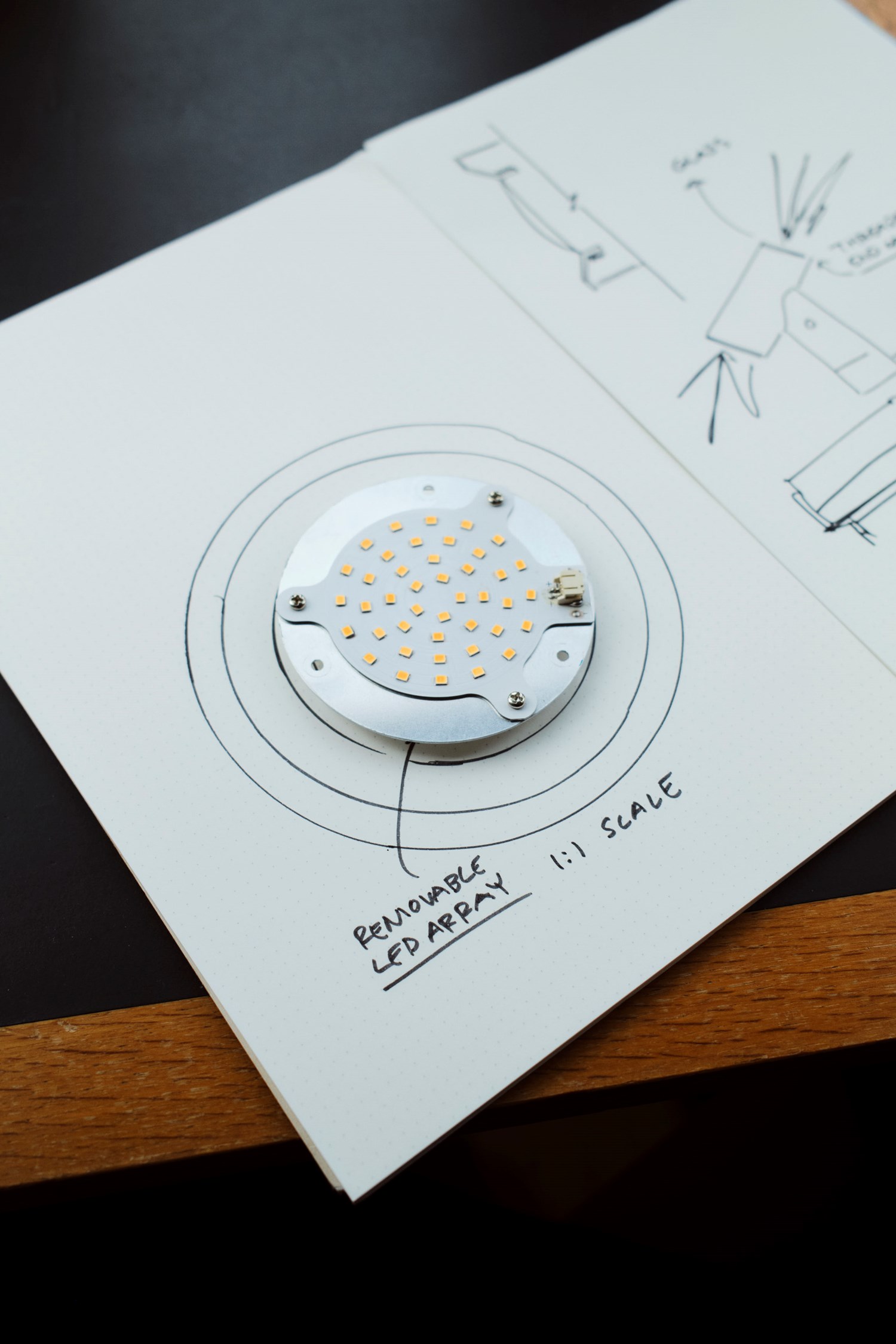 An open notebook with hand-drawn sketches and notes on lighting design rests on a dark table with wooden trim. A circular LED component sits on the page, aligning with the technical diagrams. An open notebook with hand-drawn sketches and notes on lighting design rests on a dark table with wooden trim. A circular LED component sits on the page, aligning with the technical diagrams.