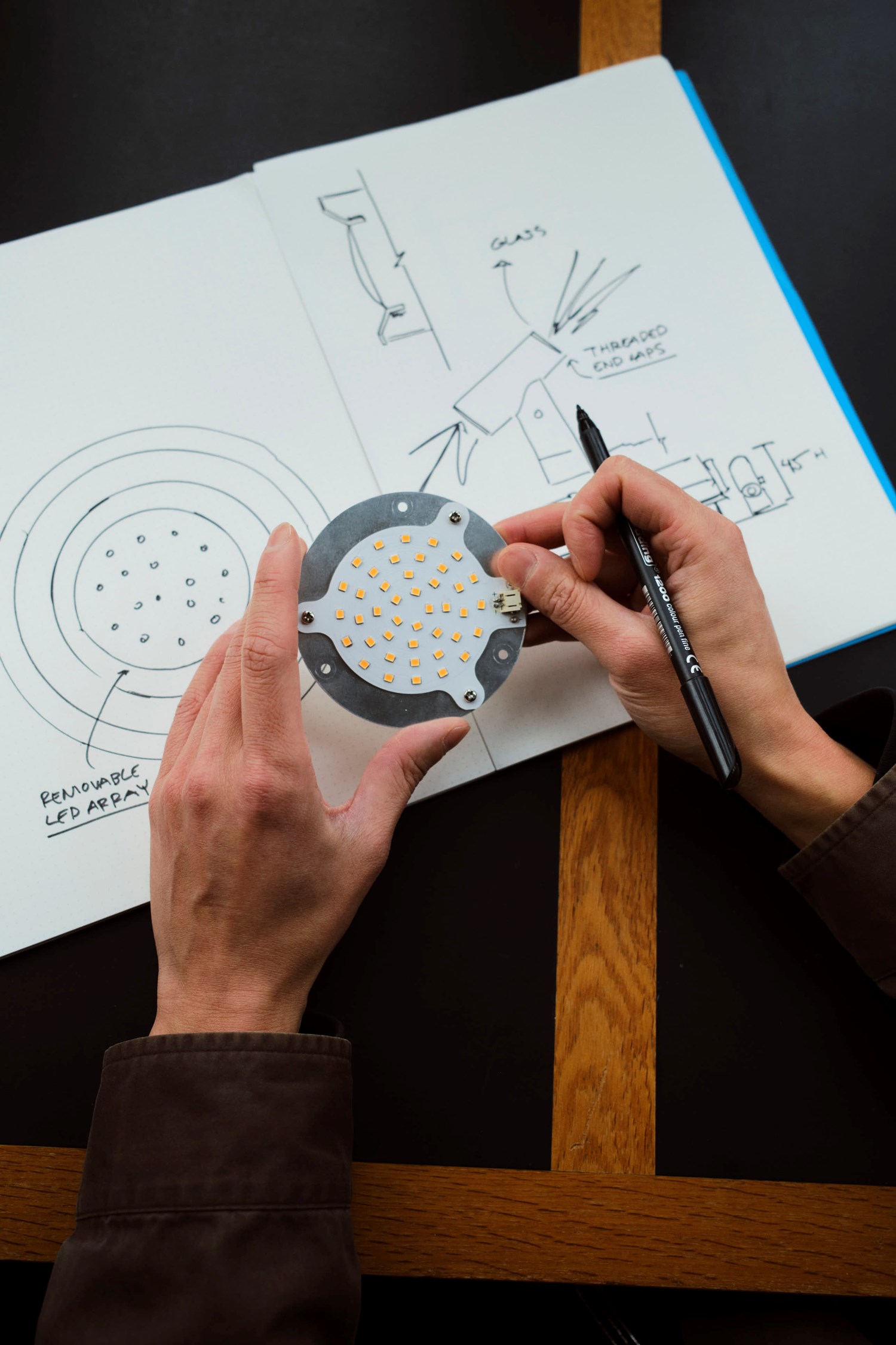 A pair of hands holds a circular LED component while sketching design ideas in a notebook. The open pages feature hand-drawn diagrams and notes. The dark table with wooden trim provides a warm, creative workspace. A pair of hands holds a circular LED component while sketching design ideas in a notebook. The open pages feature hand-drawn diagrams and notes. The dark table with wooden trim provides a warm, creative workspace.