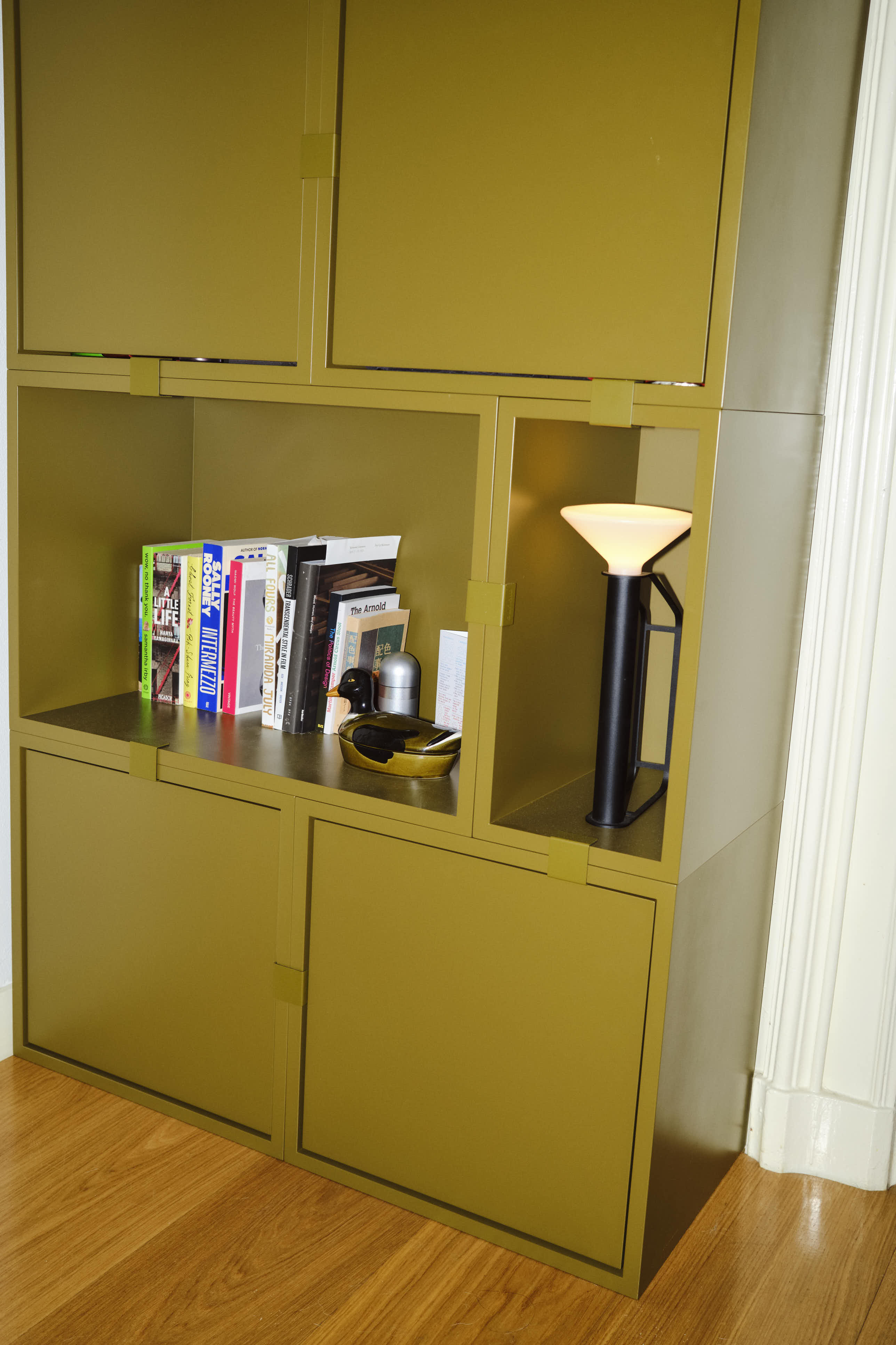 A big olive-green Stacked bookcase with books and small decorative accessories. A black Piton Portable Lamp with a white shade adds a warm glow, complementing the modern, minimalistic design.
