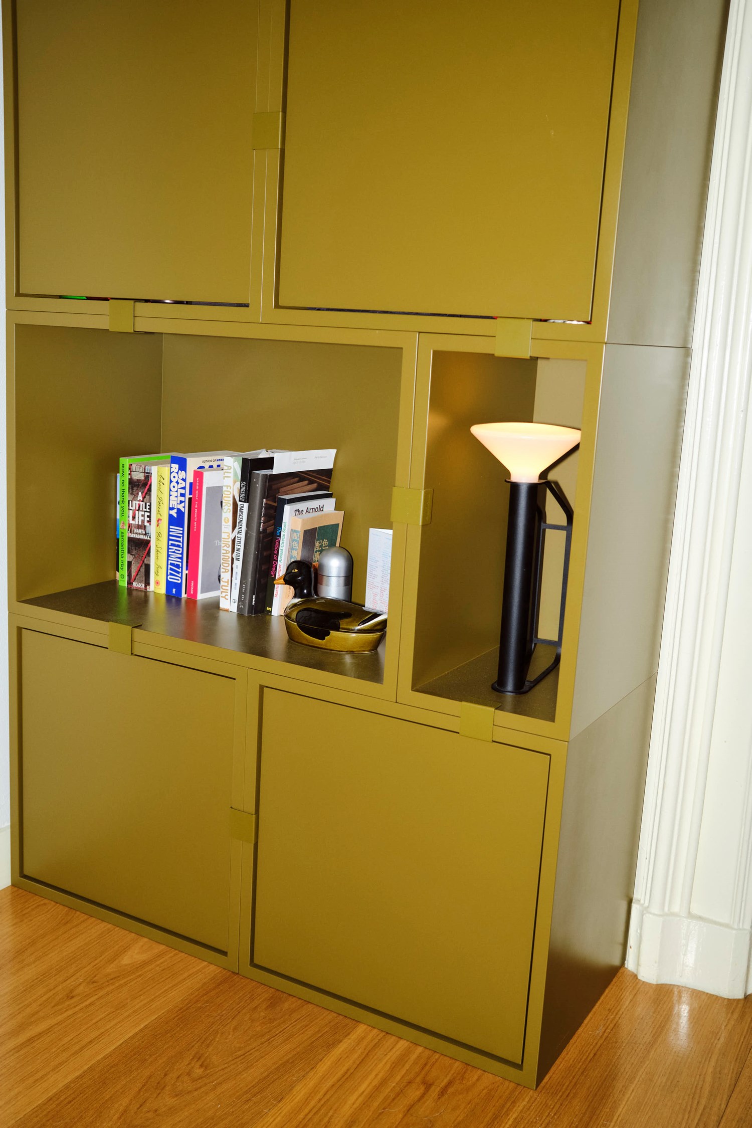 A big olive-green Stacked bookcase with books and small decorative accessories. A black Piton Portable Lamp with a white shade adds a warm glow, complementing the modern, minimalistic design. A big olive-green Stacked bookcase with books and small decorative accessories. A black Piton Portable Lamp with a white shade adds a warm glow, complementing the modern, minimalistic design.