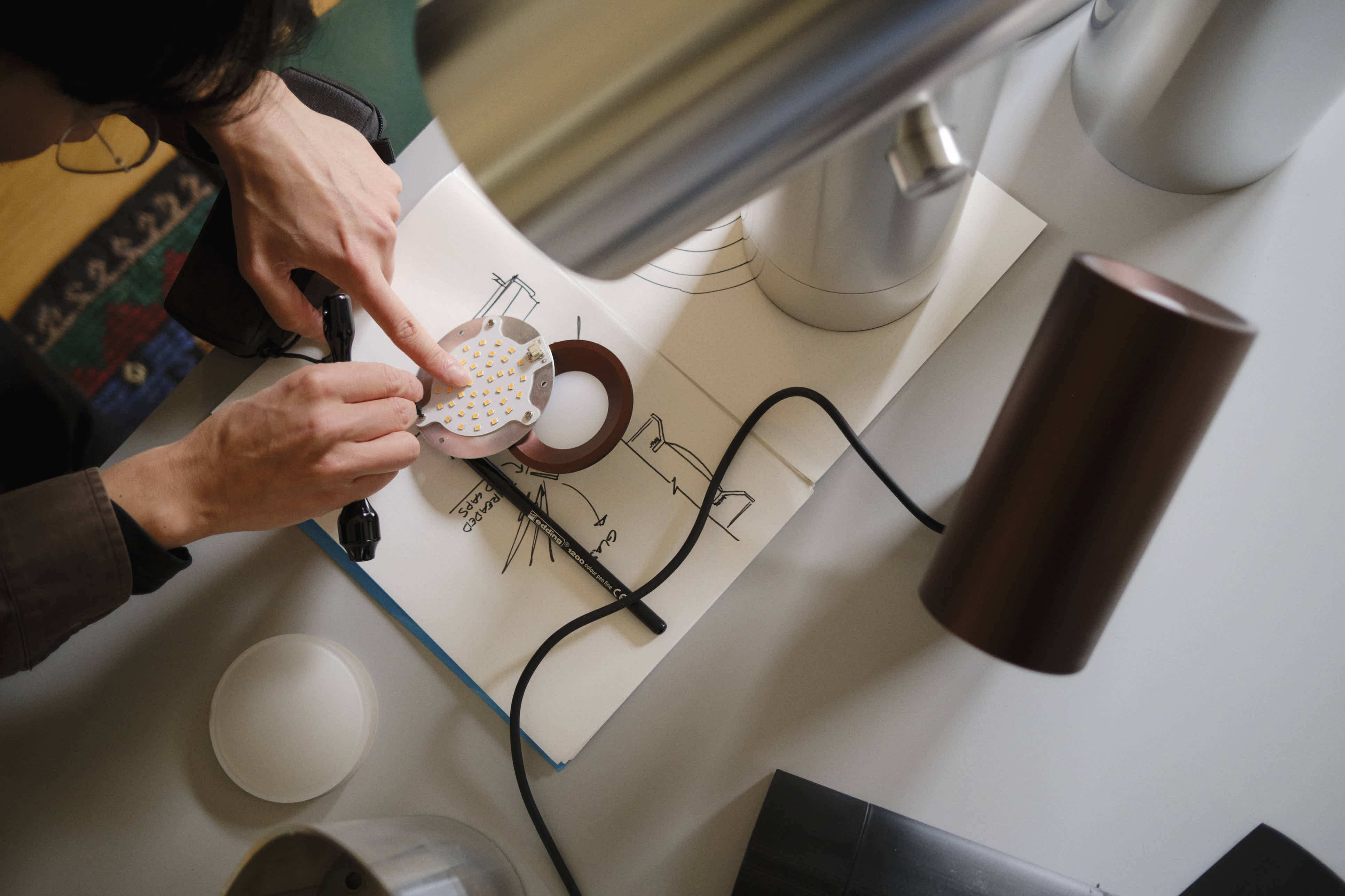 A close-up of Tom Chung working on a lighting prototype. An open sketchbook with technical drawings lies on the table, with LED components placed on top. A Beam Table Lamp and other lighting elements are placed on the table, reflecting a workspace dedicated to product design.