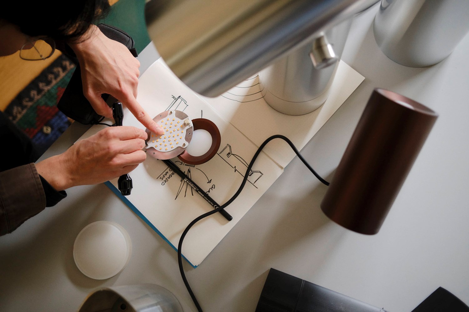 A close-up of Tom Chung working on a lighting prototype. An open sketchbook with technical drawings lies on the table, with LED components placed on top. A Beam Table Lamp and other lighting elements are placed on the table, reflecting a workspace dedicated to product design. A close-up of Tom Chung working on a lighting prototype. An open sketchbook with technical drawings lies on the table, with LED components placed on top. A Beam Table Lamp and other lighting elements are placed on the table, reflecting a workspace dedicated to product design.
