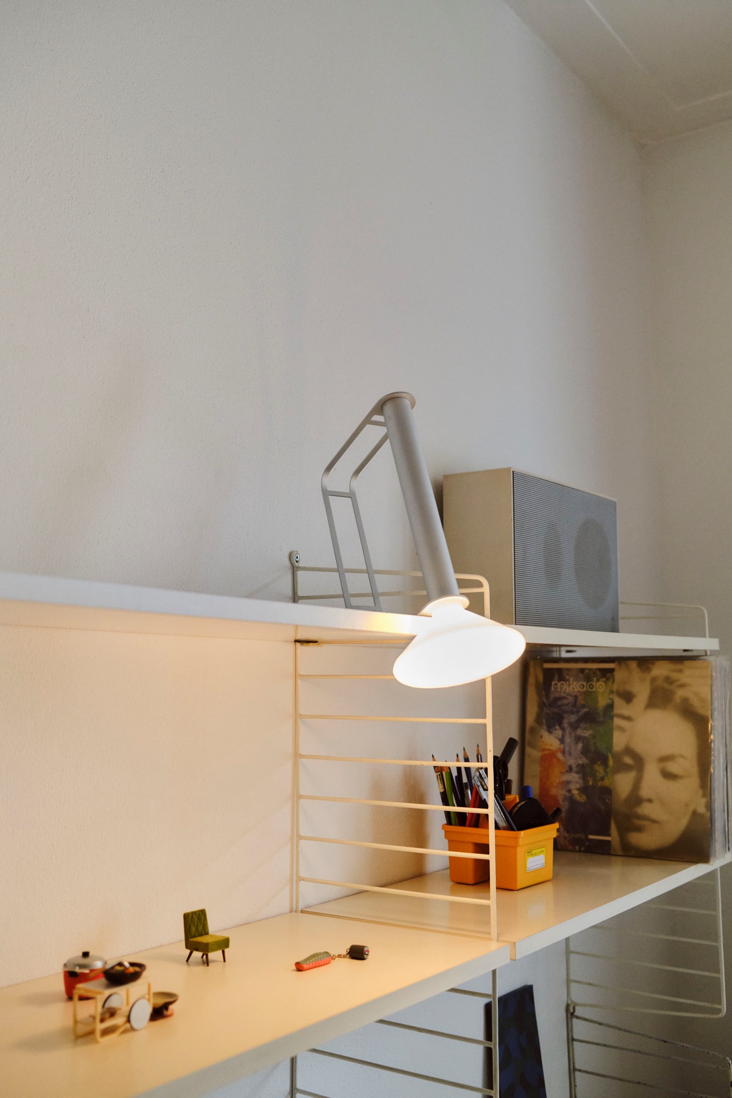 A modern white metal shelf featuring a silver Piton Portable Lamp casting a warm glow. The shelf holds small decorative items, a container of pens, and a collection of vinyl records, creating a stylish and functional workspace. A modern white metal shelf featuring a silver Piton Portable Lamp casting a warm glow. The shelf holds small decorative items, a container of pens, and a collection of vinyl records, creating a stylish and functional workspace.