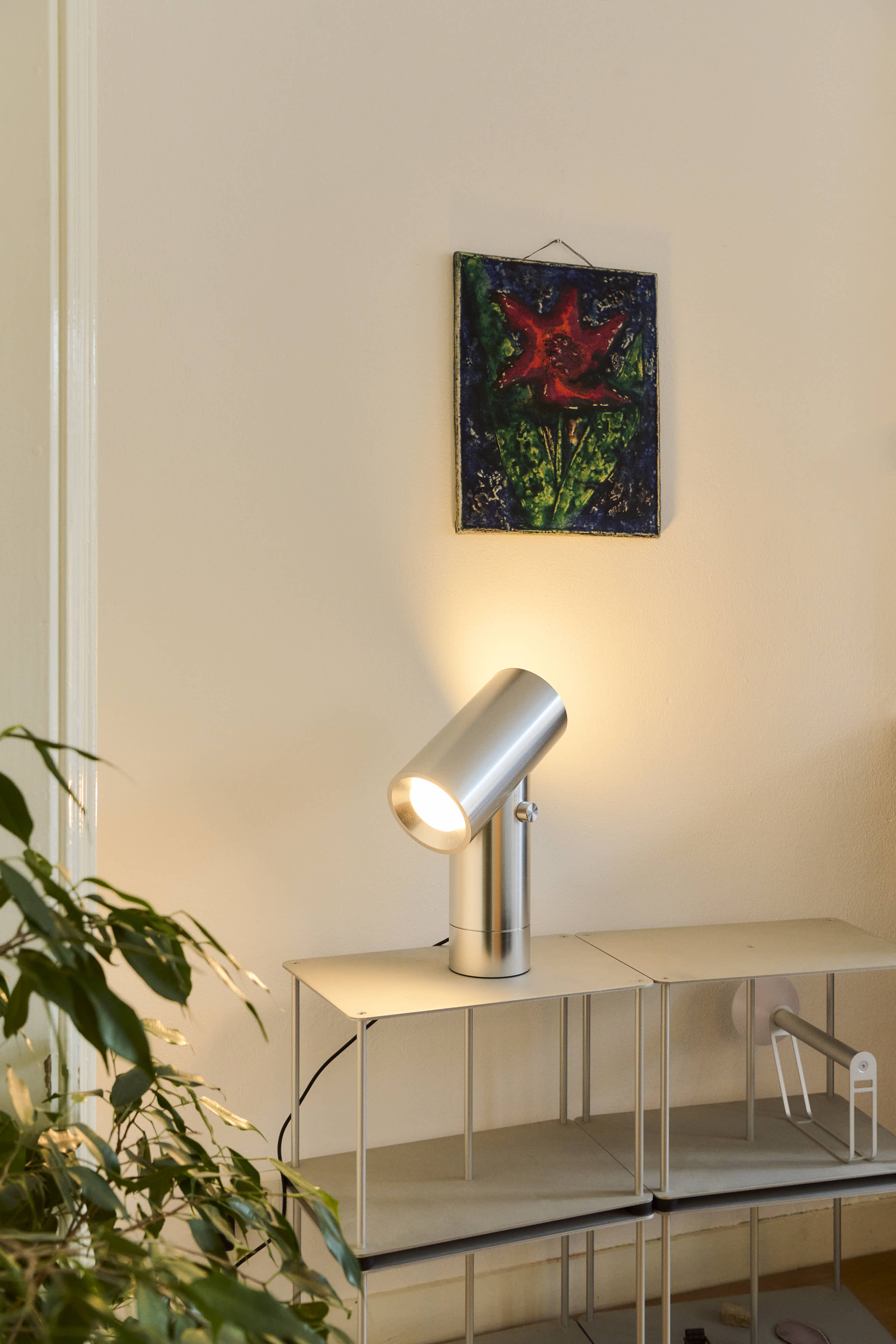 A modern interior featuring a metal shelf with a silver Beam Table Lamp and decorative objects. A red flower painting adds a striking contrast against the white wall, while a green plant peeks into the frame.