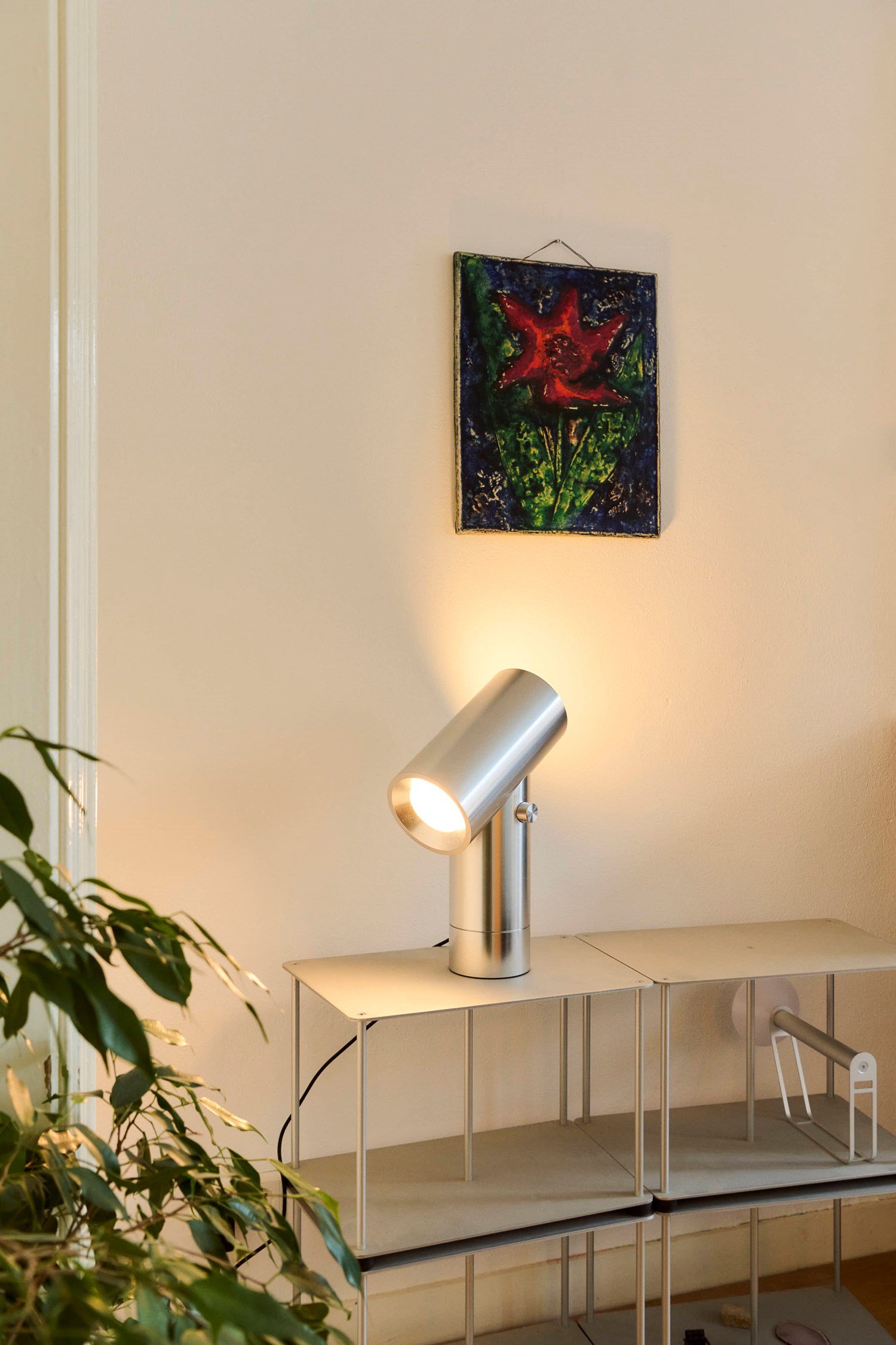 A modern interior featuring a metal shelf with a silver Beam Table Lamp and decorative objects. A red flower painting adds a striking contrast against the white wall, while a green plant peeks into the frame. A modern interior featuring a metal shelf with a silver Beam Table Lamp and decorative objects. A red flower painting adds a striking contrast against the white wall, while a green plant peeks into the frame.