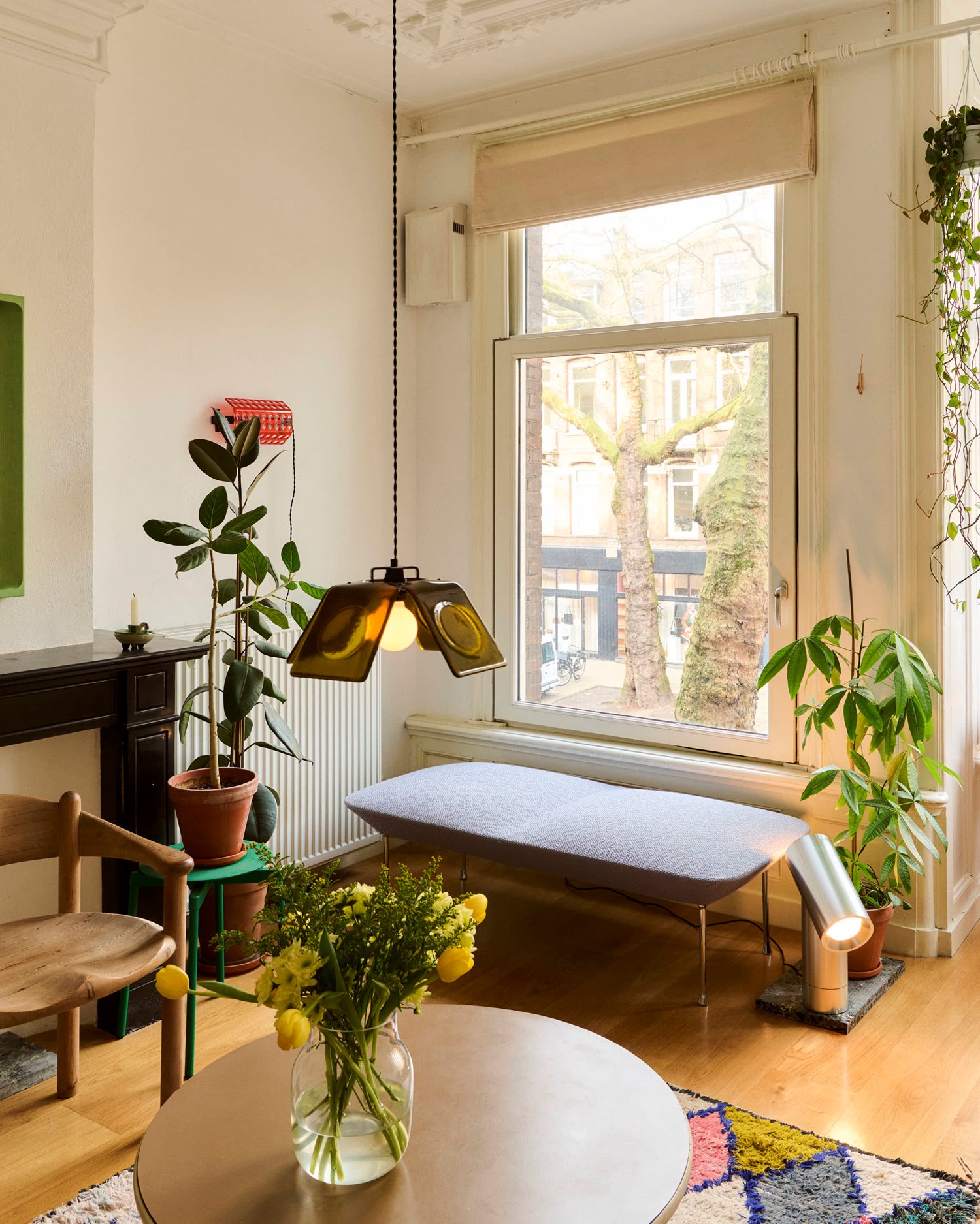 A cozy living space with a large window, green plants, and a light grey bench. A round table holds yellow flowers, while a pendant and Beam Table Lamp add warm lighting.  A cozy living space with a large window, green plants, and a light grey bench. A round table holds yellow flowers, while a pendant and Beam Table Lamp add warm lighting.