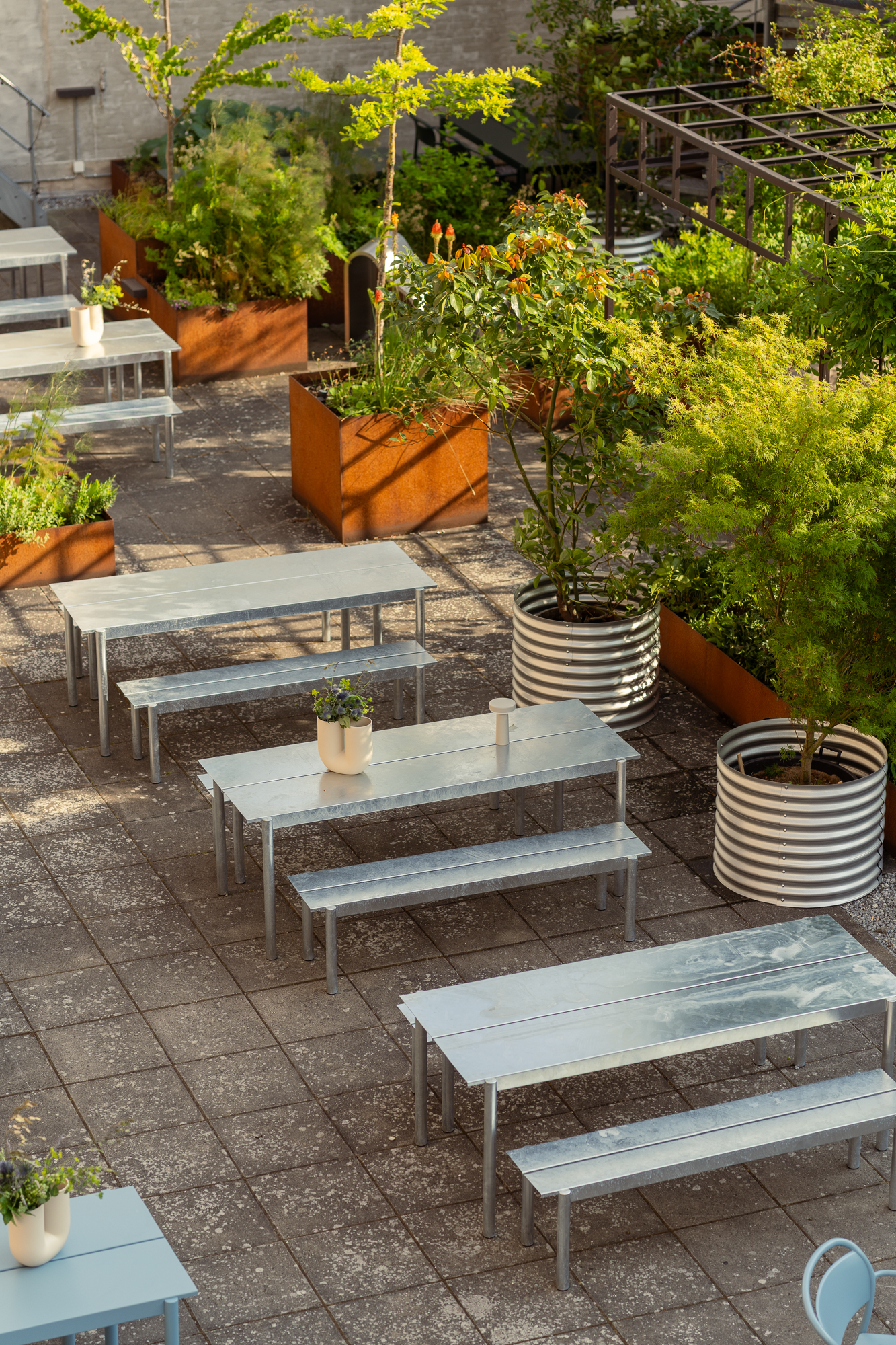 Linear Steel Tables and benches are placed in a sunny courtyard surrounded by green plants and vases with colorful flowers. 