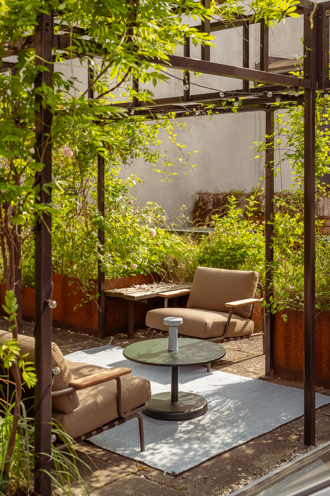 A Settle sofa and matching chair are arranged in a sunny courtyard, surrounded by lush green plants. The seating ensemble rests on a light beige rug, complemented by an outdoor table, creating a cozy and inviting atmosphere.