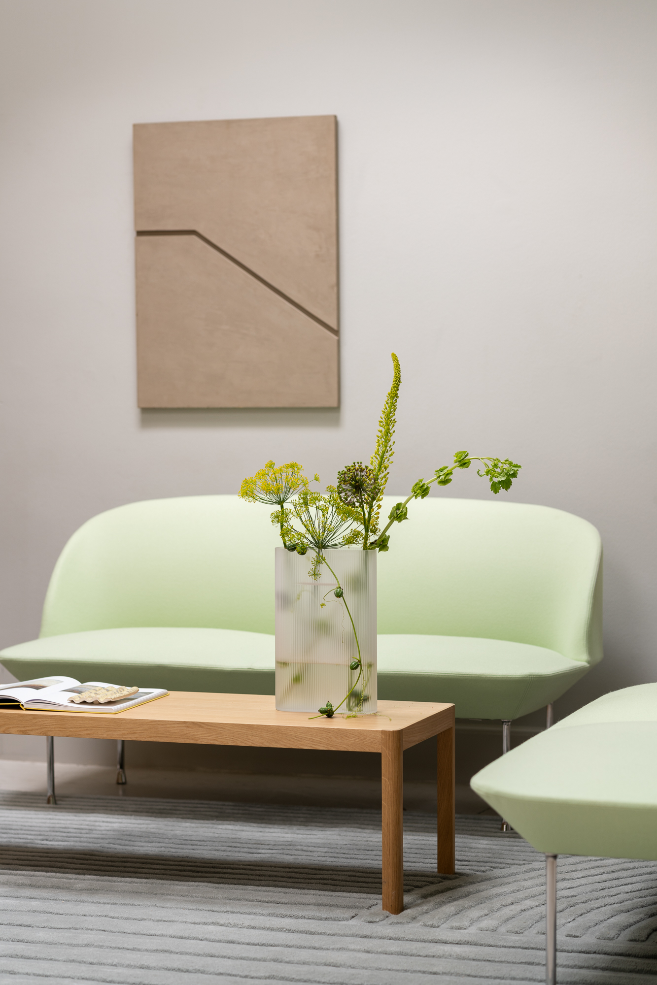 The Oslo Sofa in light green is placed behind a minimalist wooden coffee table. An artistic, minimalistic vase with green flowers sits on the table, adding a cozy and inviting touch to the room.
