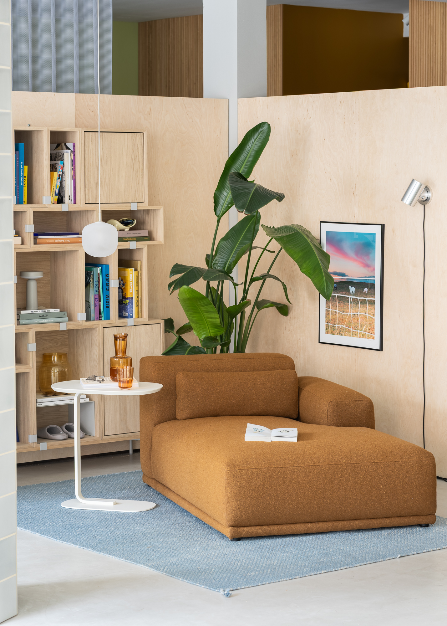 A brown Connect Sofa is positioned on a light blue rug, styled with a white sofa table that adds contrast and brightness to the space. A large green plant brings a fresh, natural element to the room, complemented by a wooden storage system that adds warmth and functionality.