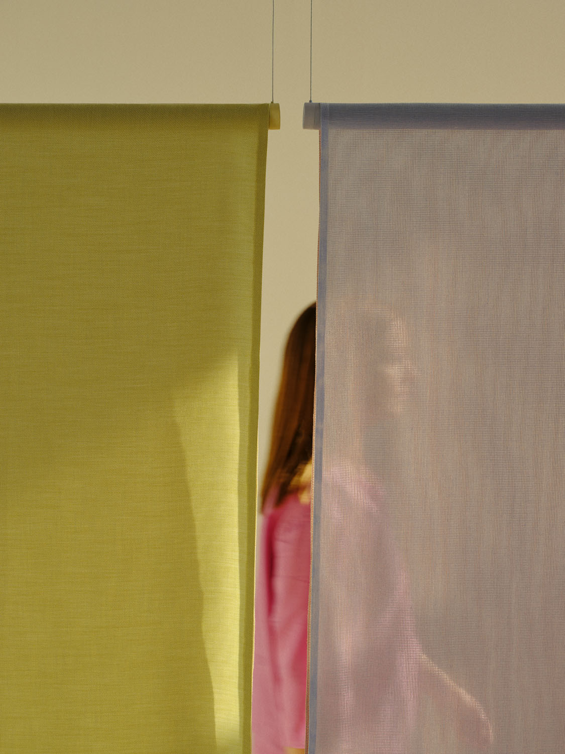 Two sheer textile panels from the “Veil” collection—one in citrus green and the other in dusty lavender—are suspended side by side. Behind them, a figure in a pink top is faintly visible through the mesh, demonstrating the screen’s layered transparency and spatial division effect.