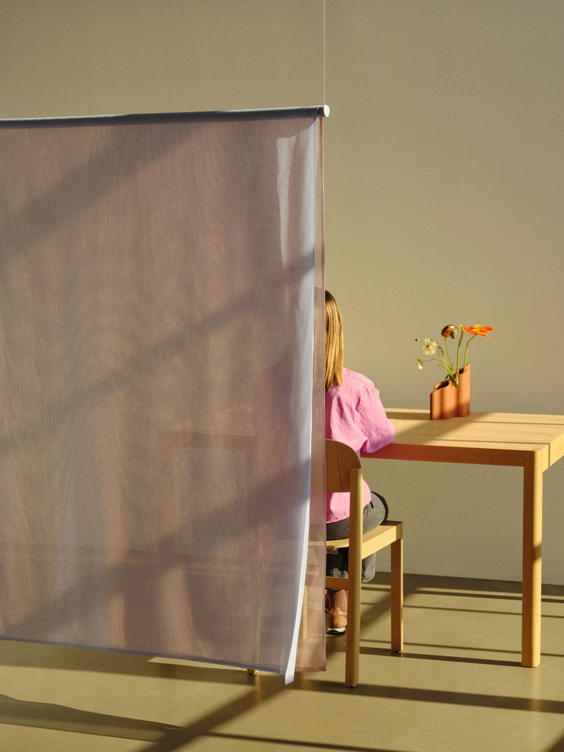 A warm-toned interior scene showing a person seated at a wooden table, partially obscured by a suspended semi-transparent fabric panel. The panel, part of the "Veil" project by Nikola & Florian, creates a soft division in space.

